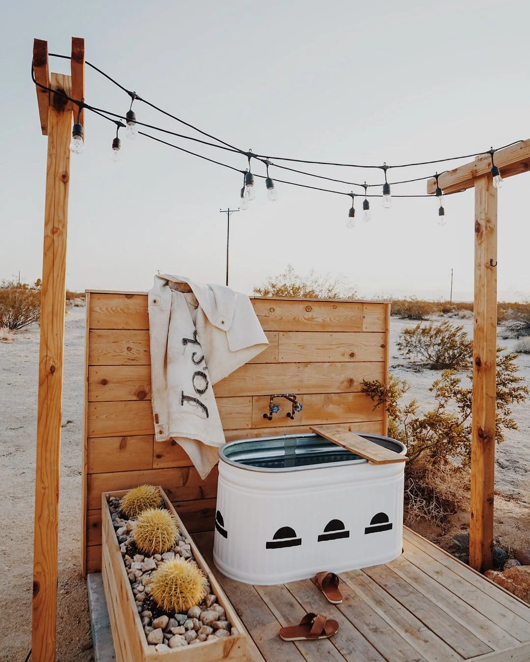 Outdoor shower setup with wooden frame, string lights, bath tub, towel, and potted cacti in a desert landscape.