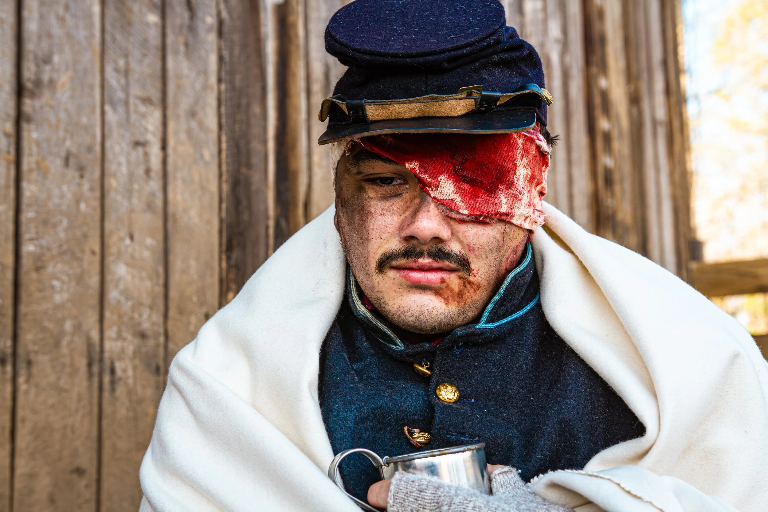 A young man with dirt on his face wearing a black hat with goggles, a red cloth over his right eye, and a white blanket draped over his shoulders, sitting against a wooden fence holding a metal cup.