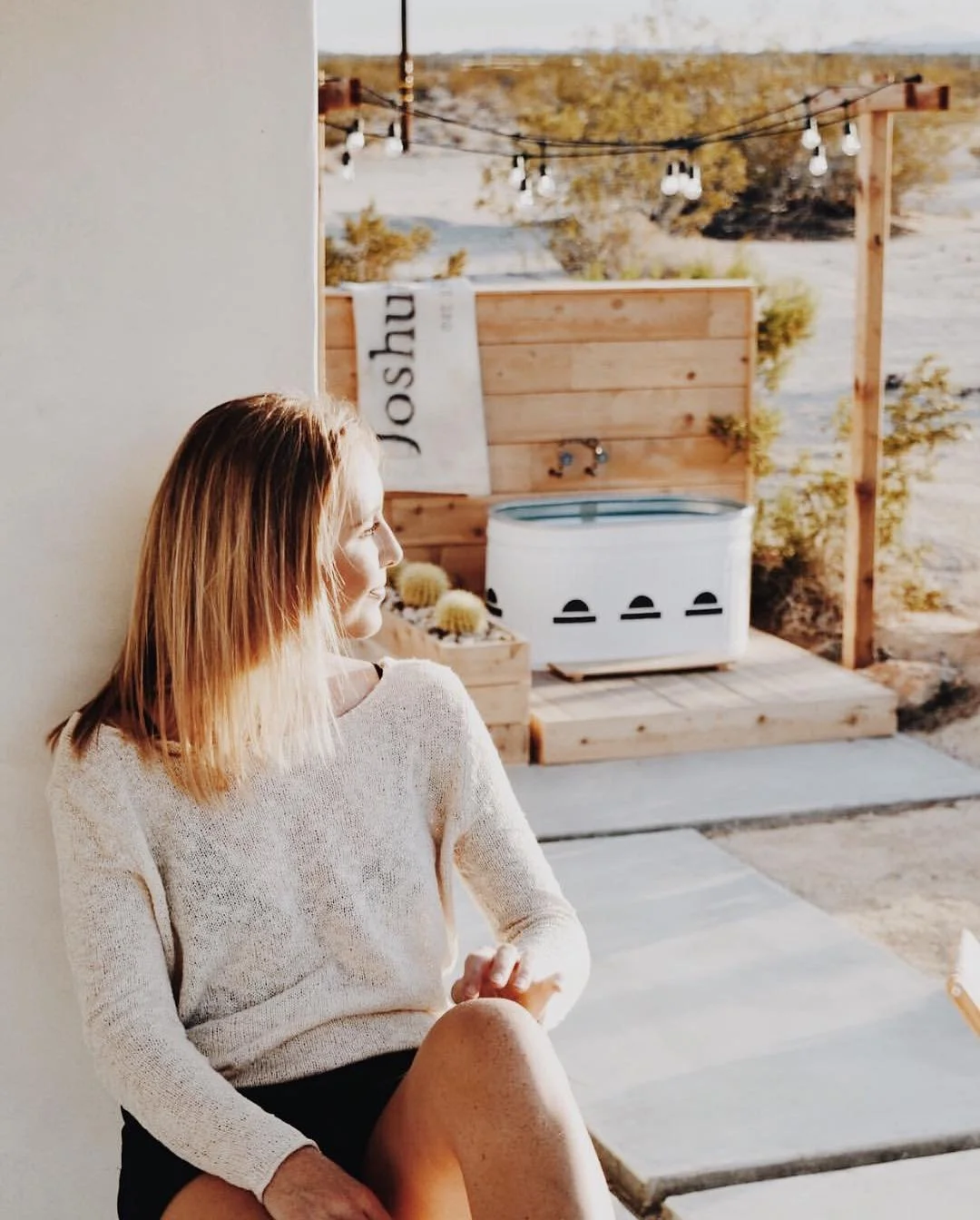 Woman with red hair sitting on a concrete patio, looking outside in sunlight, with a wooden hot tub and string lights in a desert landscape in the background.