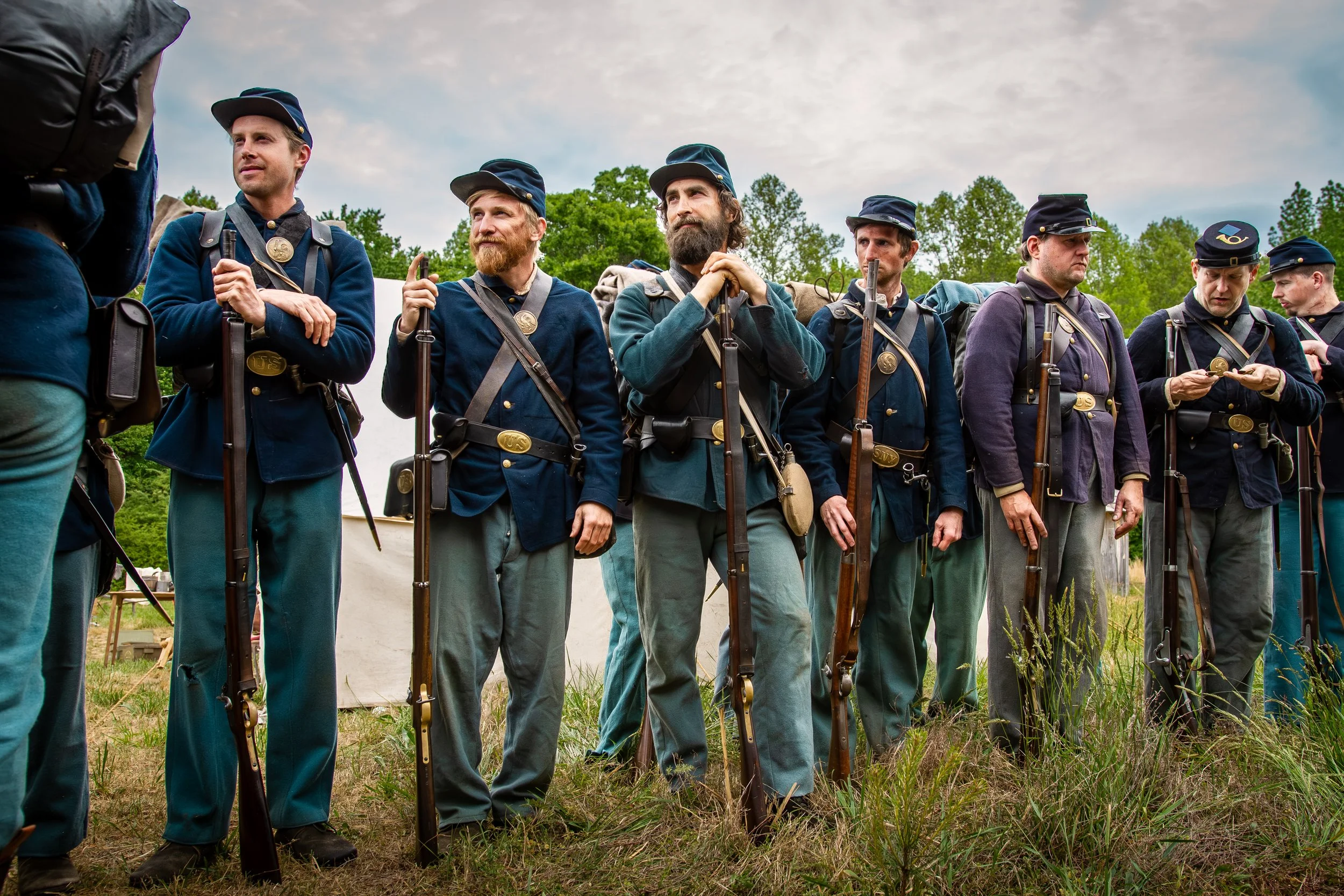 Historical reenactors dressed as 19th-century soldiers standing in line outdoors, holding rifles, with green trees and cloudy sky in the background.