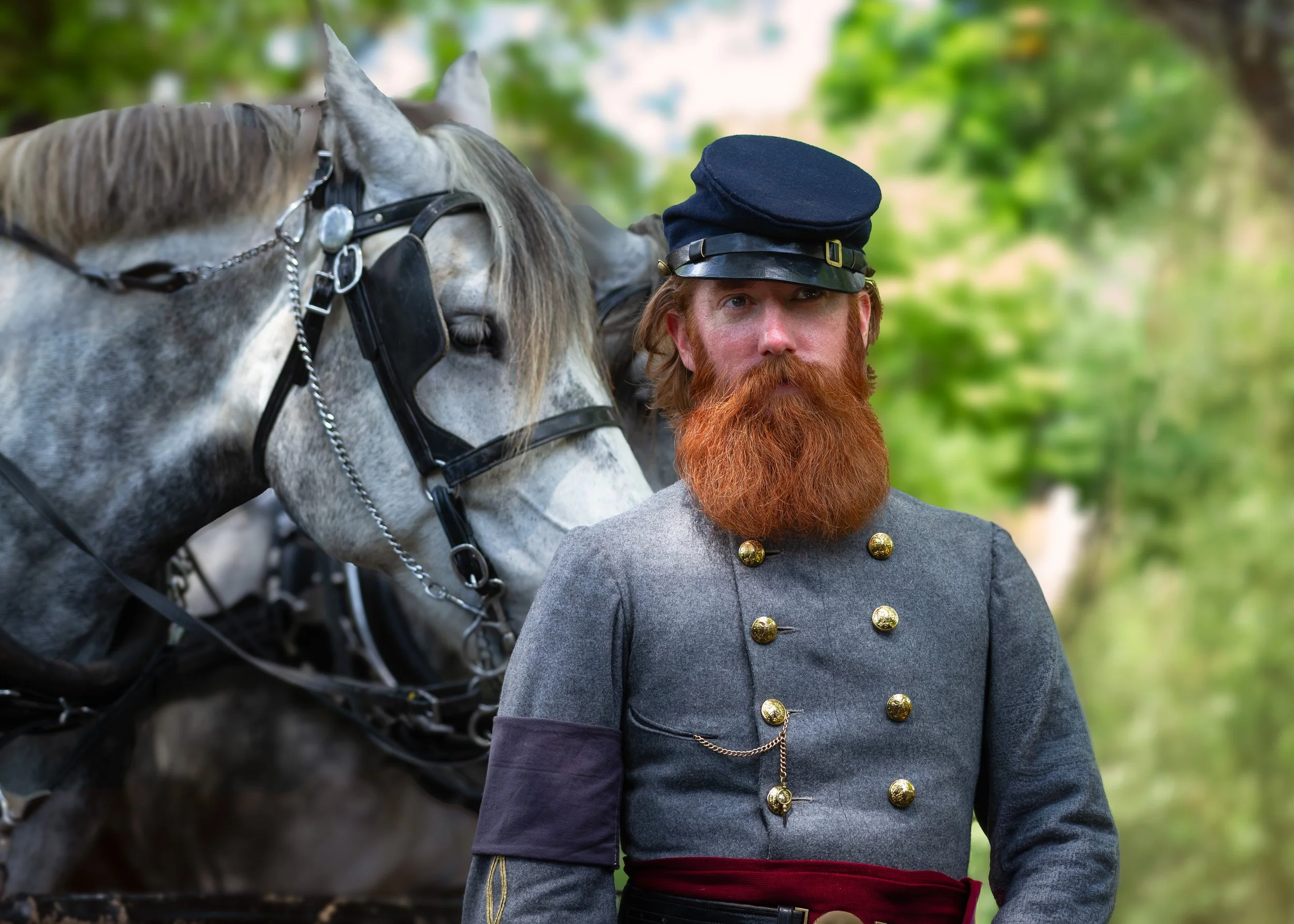 A man with a red beard wearing a gray military-style coat with gold buttons, a chain, and a dark hat, standing outdoors next to a gray horse with a black harness, with green trees in the background.