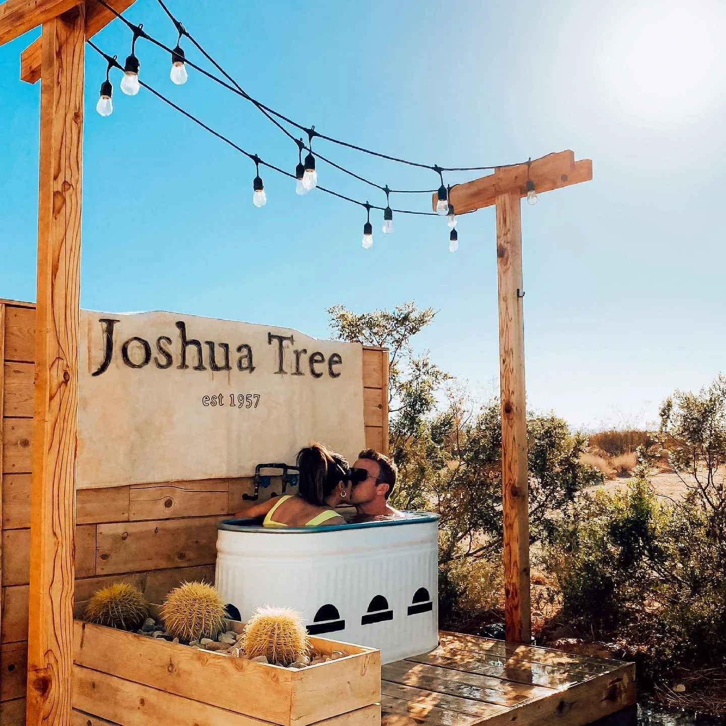 Couple kissing in a small outdoor bathtub at Shack Attack, with a flagn reading 'Joshua Tree est 1957', wood fencing, string lights, and desert vegetation in the background on a sunny day.