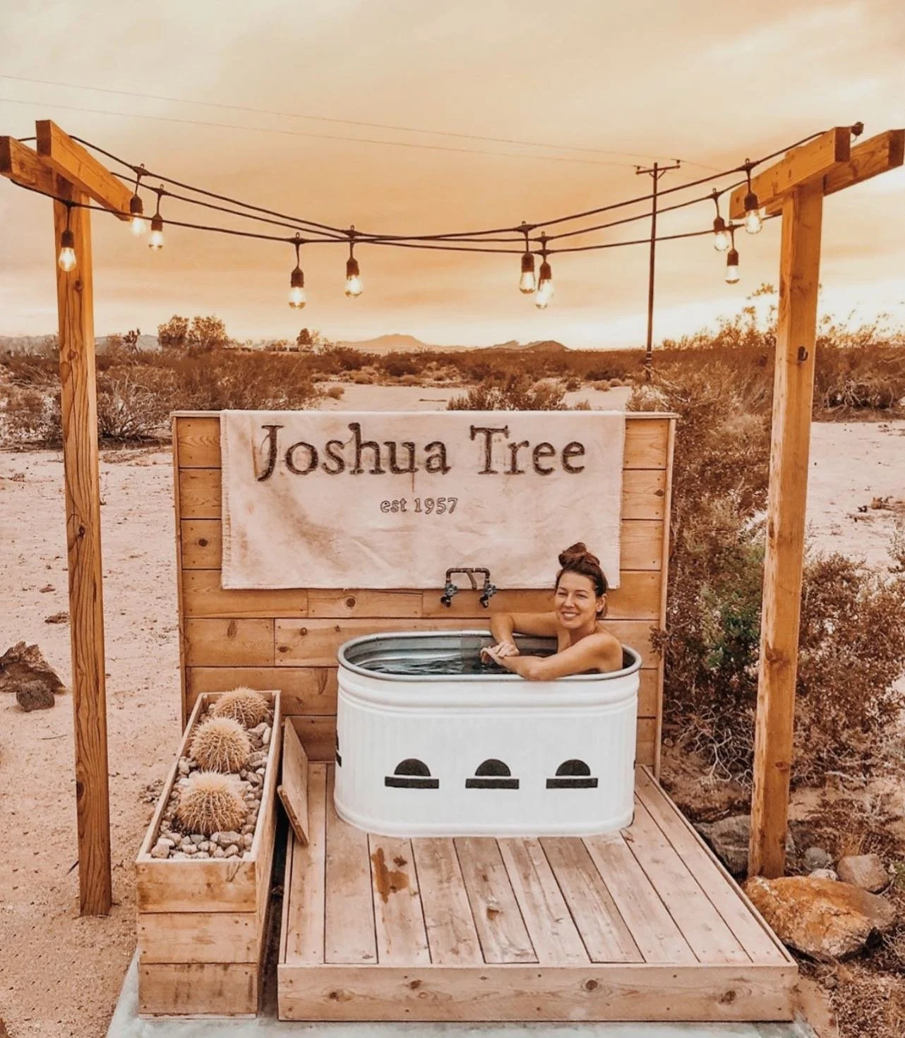 A woman relaxing in a small hot tub in a desert landscape with cacti, under a string of outdoor lights, in front of a wooden sign that reads 'Joshua Tree est 1957' and a wooden planter with cacti.