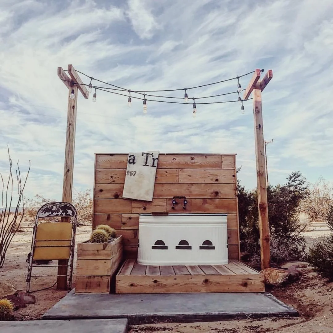 Outdoor rustic setup featuring a wooden platform with a white tub, wooden wall with a hanging sign, a vintage chair, a cactus, and string lights under a partly cloudy sky.
