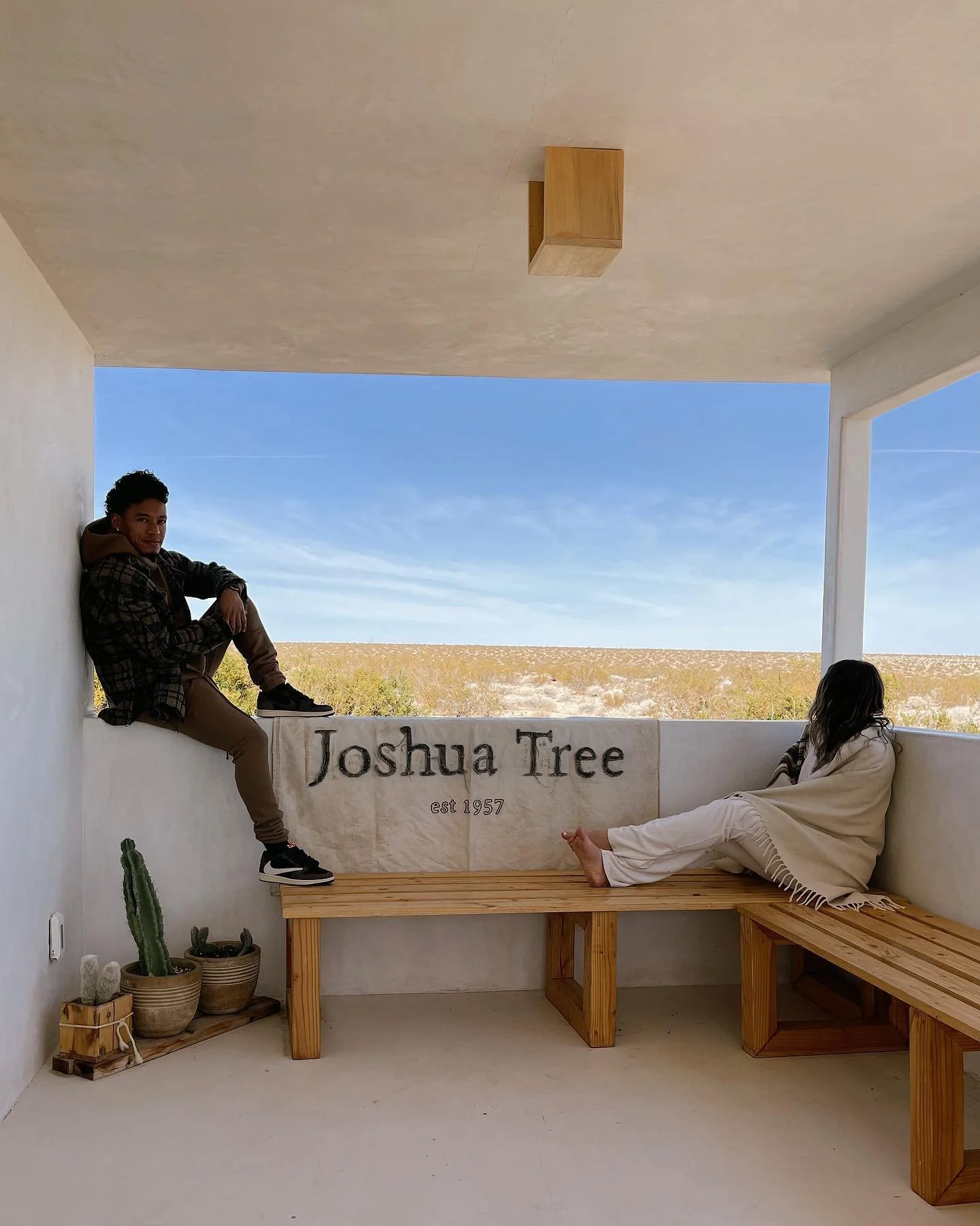 Two people sitting in a covered outdoor porch with a view of a desert landscape and blue sky. One person is sitting on a bench, and another is sitting on the wall, with a flag that says 'Joshua Tree est 1957.'