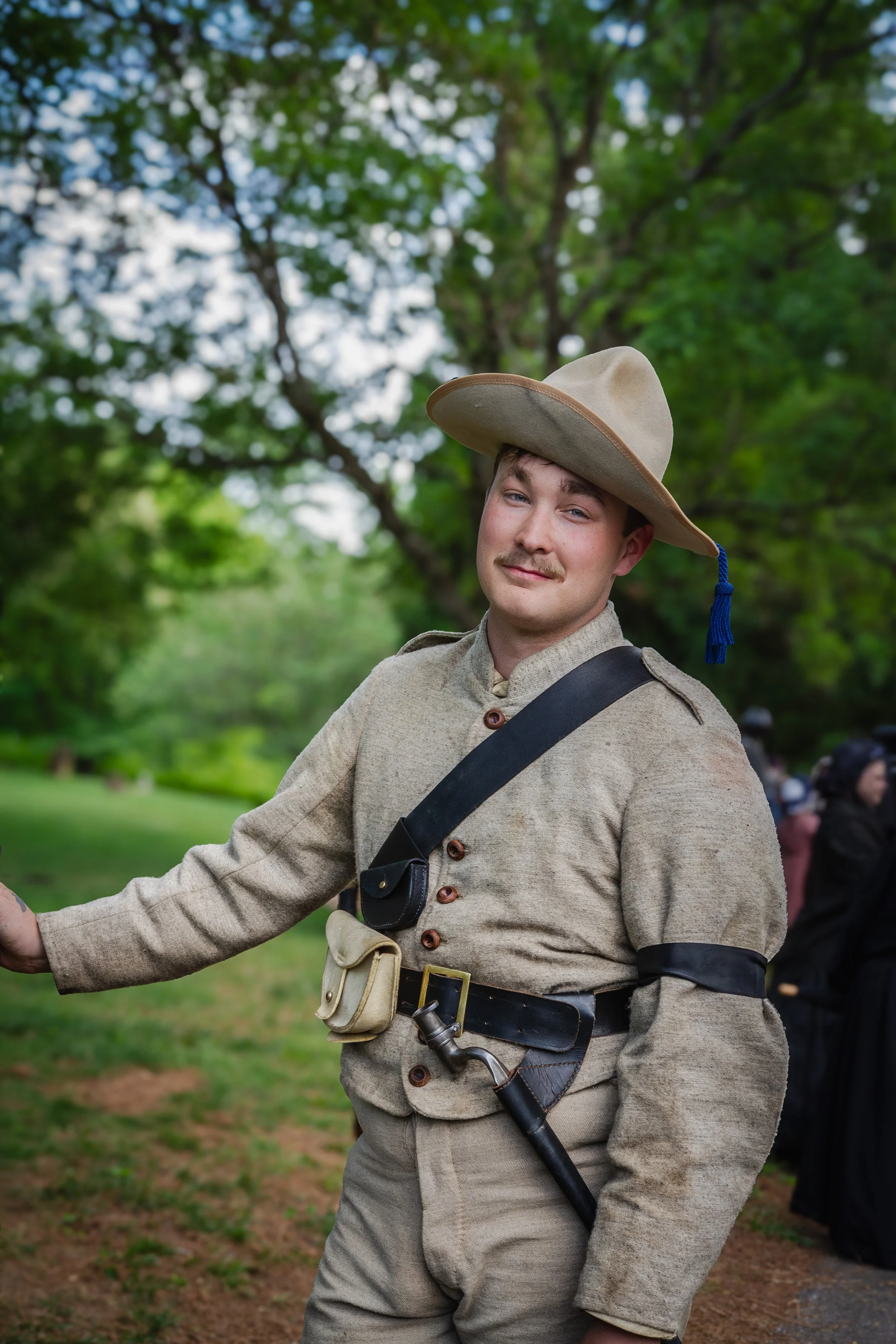 A man dressed in historical attire stands outdoors in front of green trees, wearing a wide-brimmed hat with a blue tassel, beige clothes, and gear including a sword.
