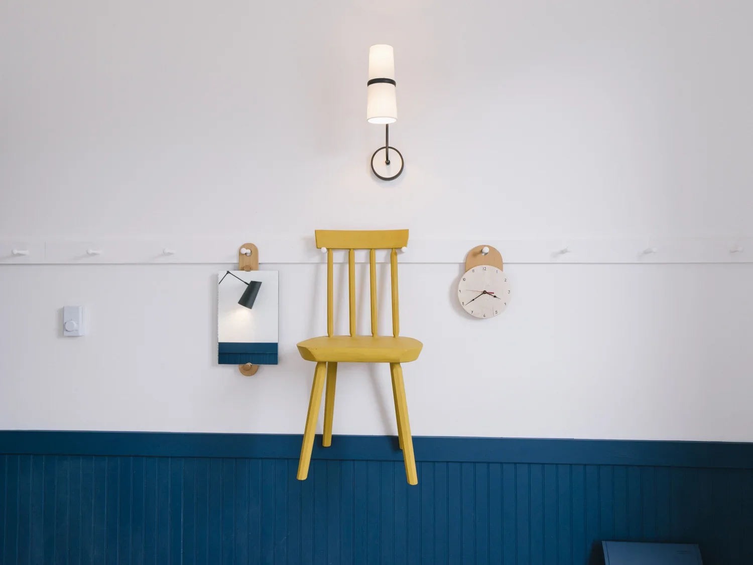 Interior wall with a yellow wooden chair mounted upside down, a small mirror, a modern clock, and a wall-mounted light fixture