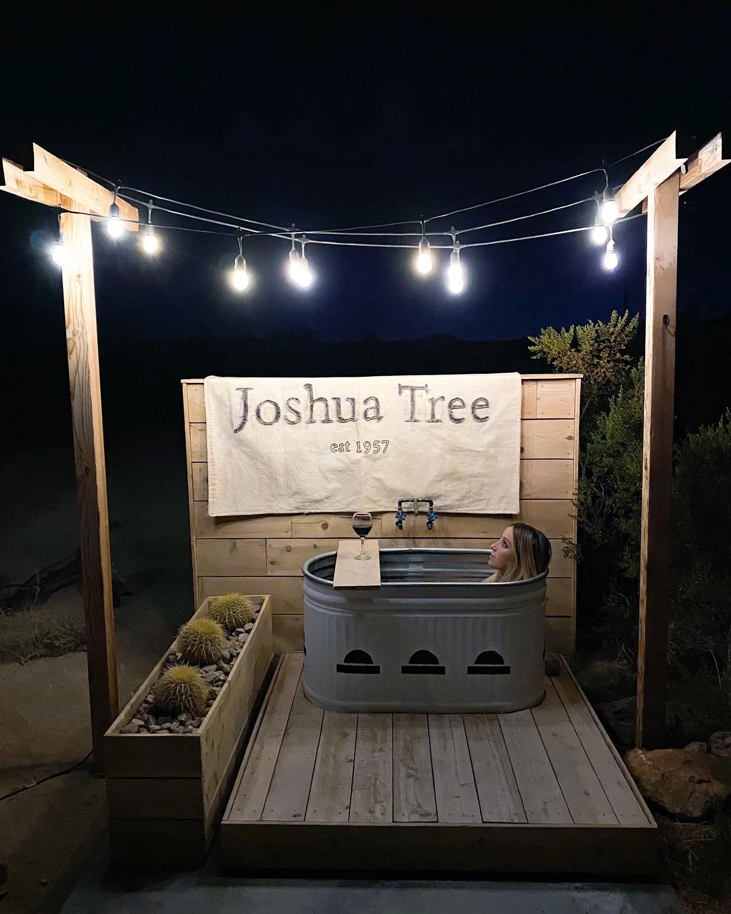 A woman soaking in a small metal hot tub on a wooden deck at night, with string lights overhead and a wooden privacy wall with a banner that reads 'Joshua Tree est 1957', a wine glass on a wooden plank, a faucet, and a planter with cacti nearby.