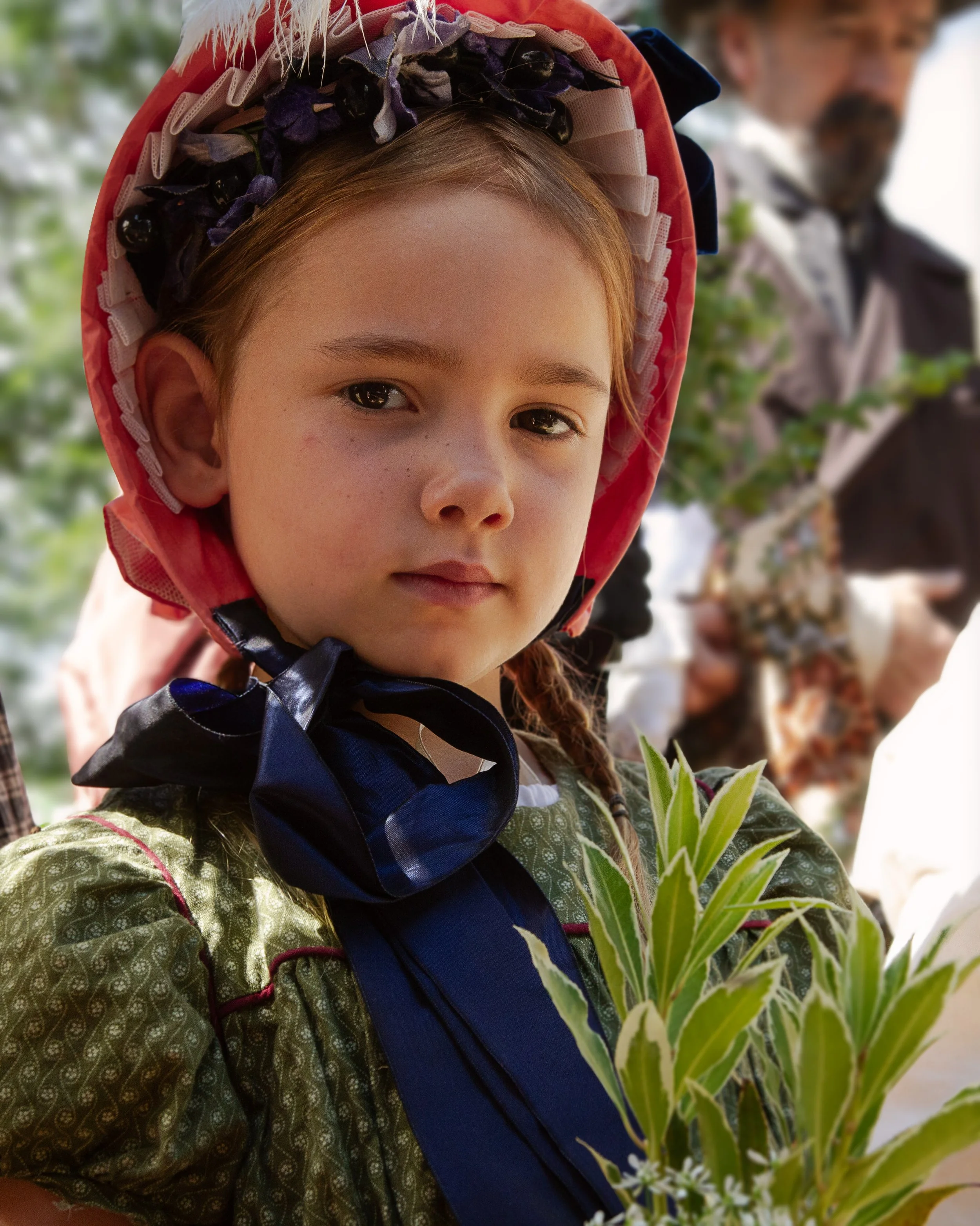 A young girl with brown hair and light skin, wearing a decorative hat and traditional attire, holding green leafy plants. Two adults are blurred in the background, one woman with red hair and a man wearing sunglasses and a vest, outdoors with trees.