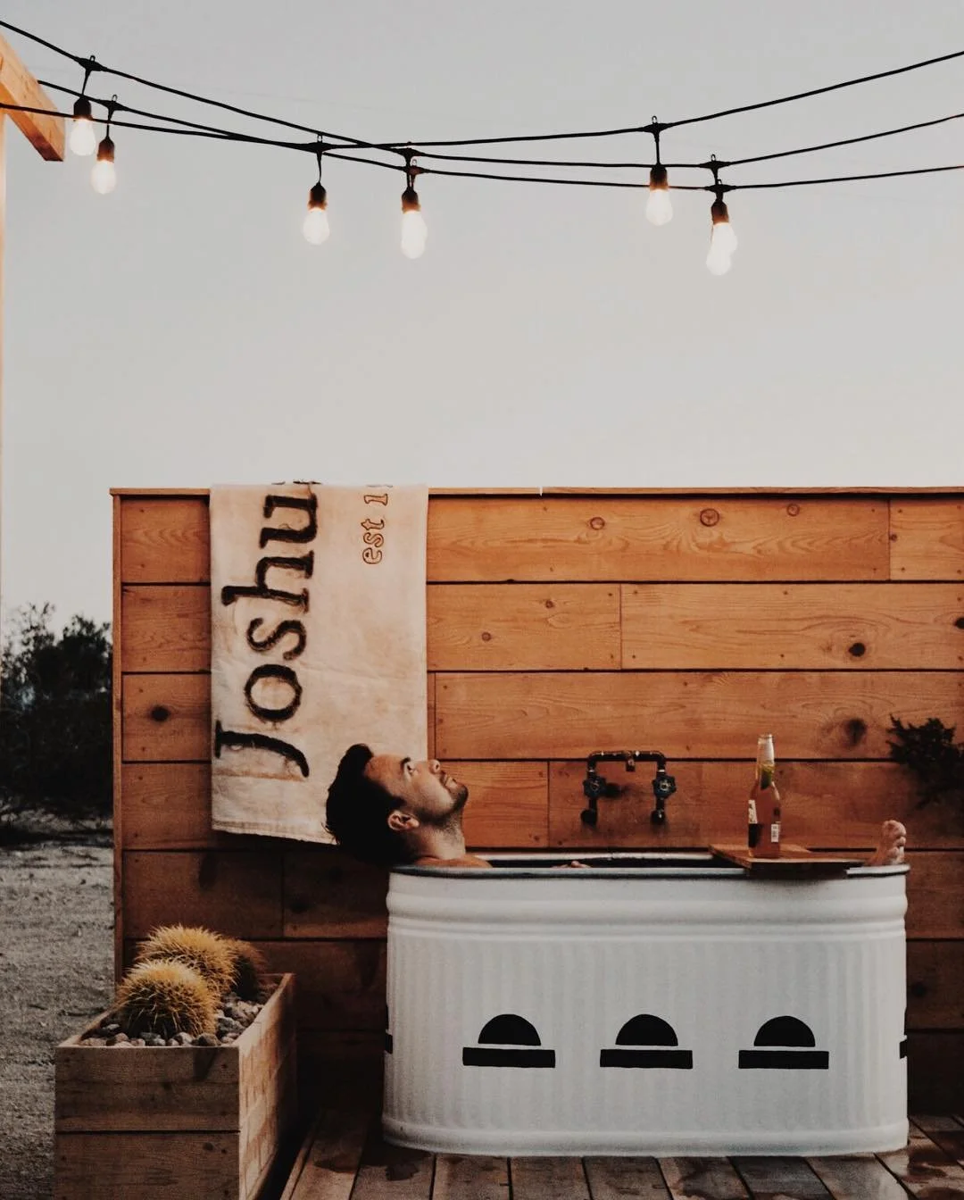 A man relaxing in a vintage bathtub outdoors at sunset, with a wooden privacy wall, potted cacti, string lights overhead, and a bottle of beer on a small table.
