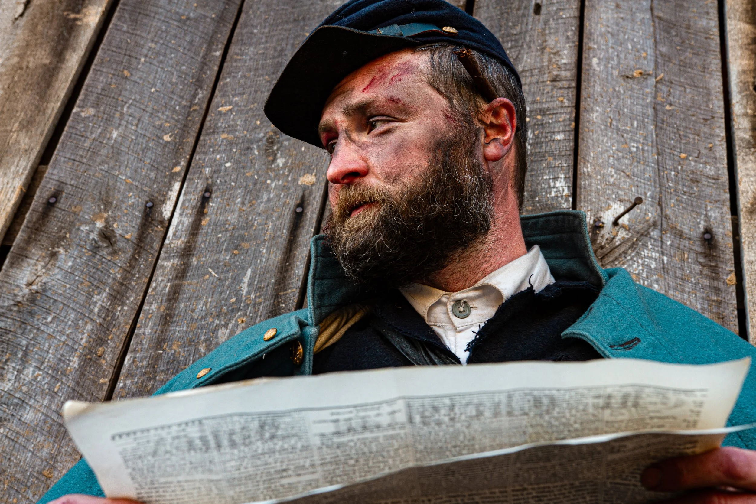 A man in historical military uniform with a beard, reading a newspaper while leaning against a wooden surface. The man has scratches on his face and is wearing a black hat and teal coat.