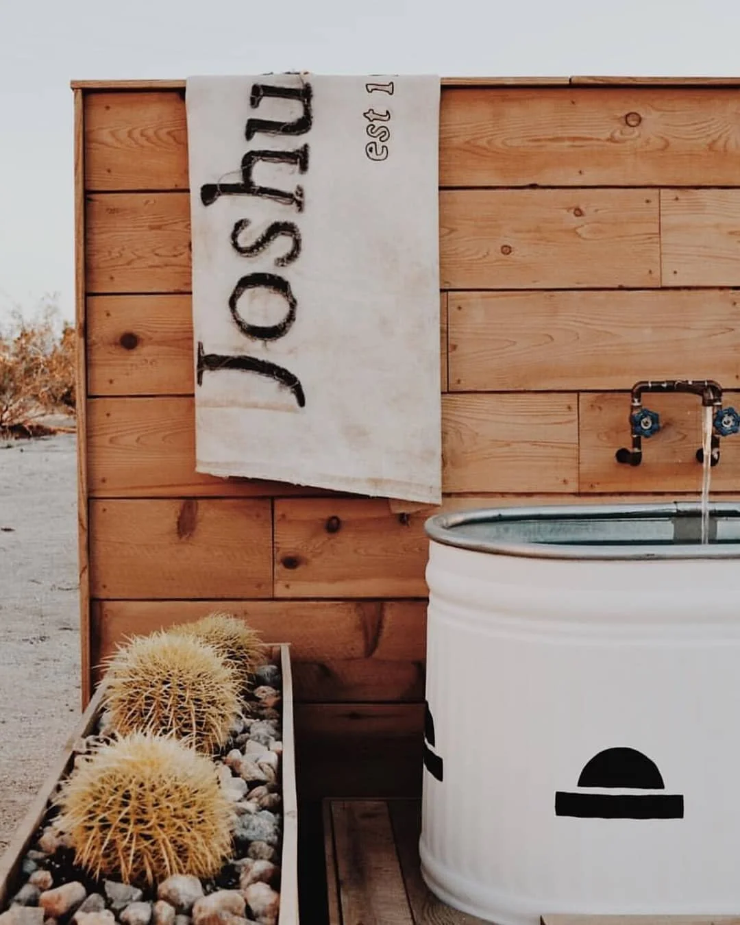 Wooden outdoor wash station with a white barrel sink, a wooden panel backdrop with a custom fabric flag that reads 'Joshua Tree' and a cactus plant in a rectangular planter.