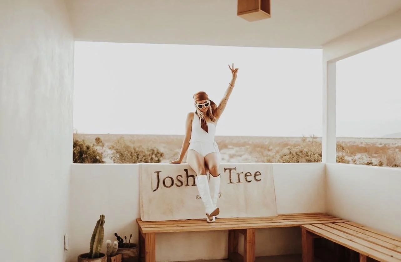 A woman in a white outfit and sunglasses sitting on a wooden bench, holding one arm up in a victory pose, with a sign behind her that reads 'Josh Tree,' on a balcony with desert landscape in the background.