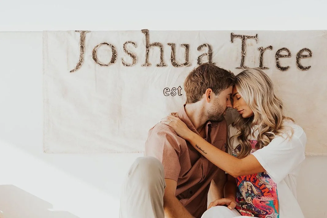 A couple sitting close together with foreheads touching, in front of a banner that reads 'Joshua Tree'.