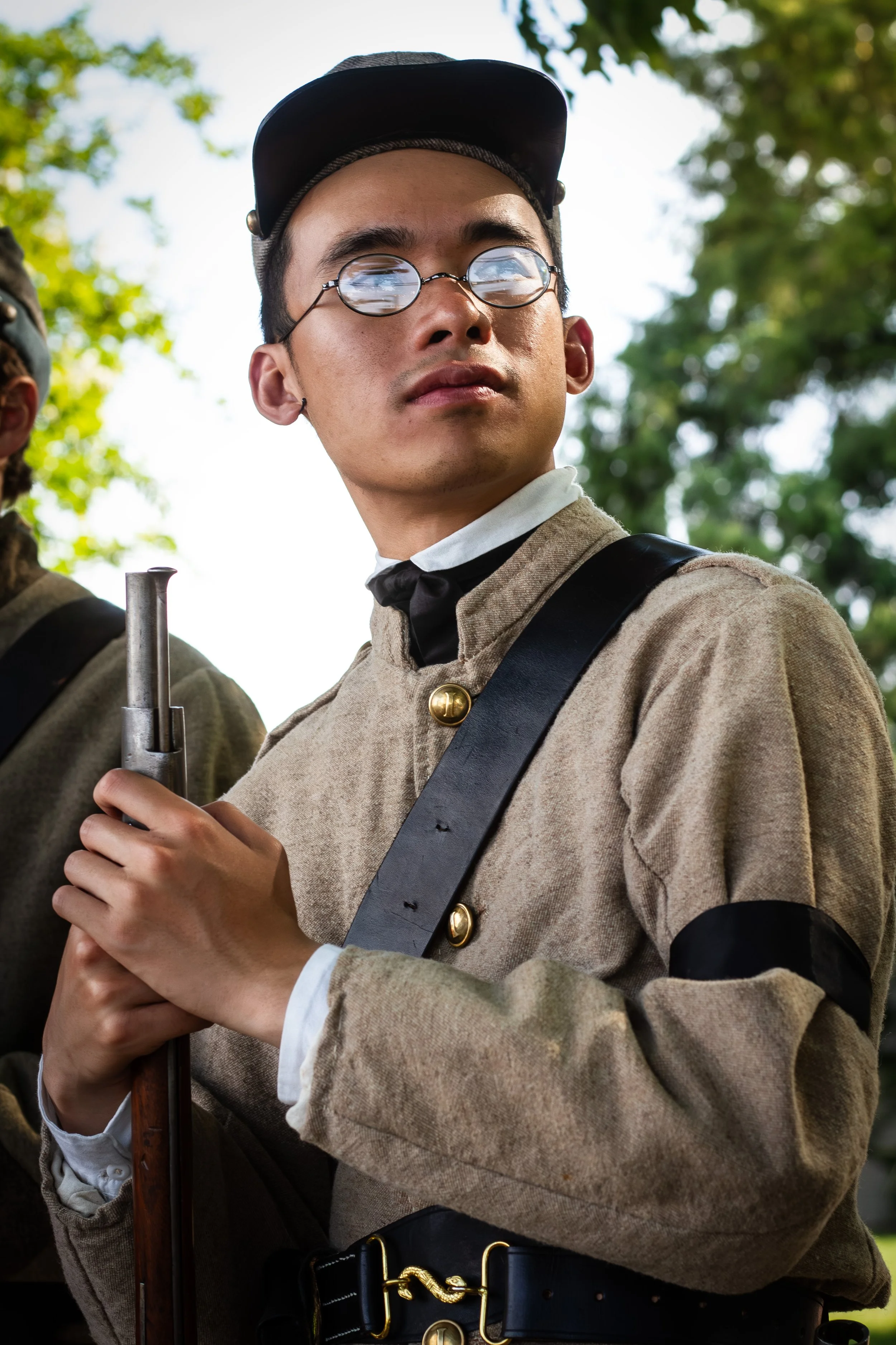 A person dressed in historical military uniform with a beige coat, white shirt, and black belt, holding a musket. They are wearing round glasses and a black cap, standing outdoors with trees in the background.