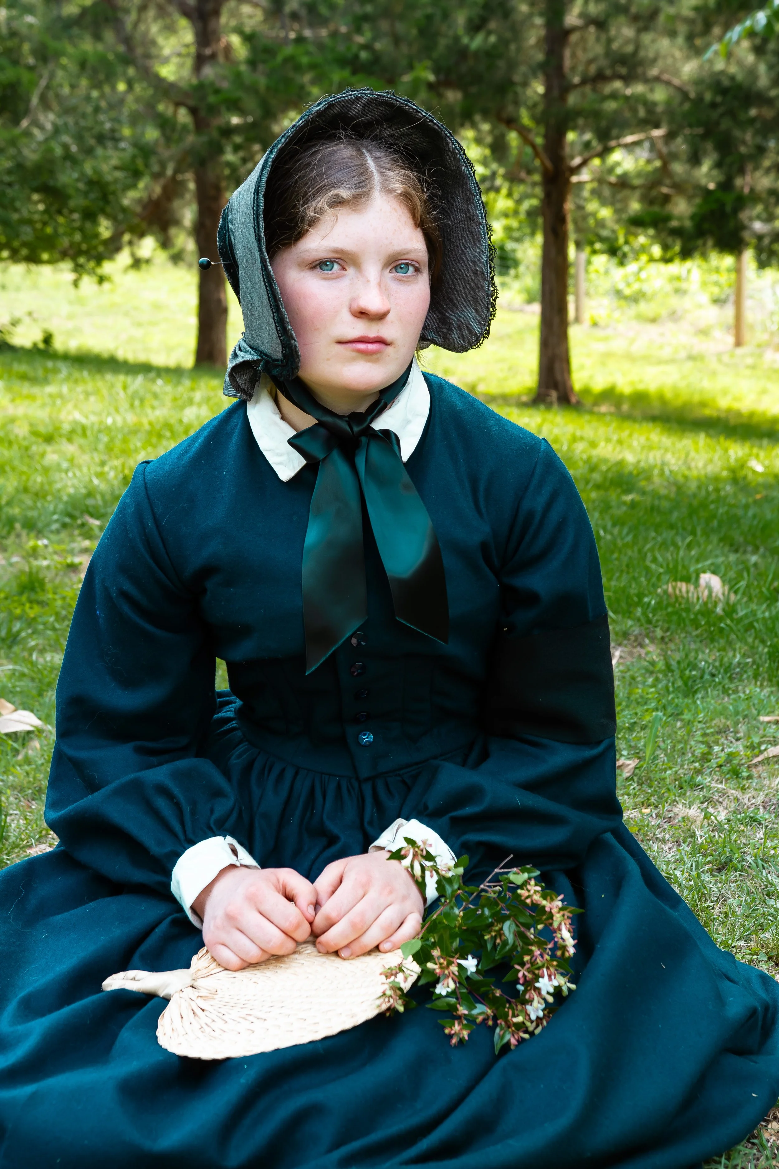 A young woman dressed in 19th-century clothing, wearing a bonnet, sitting outdoors in a grassy area with trees, holding a straw hat with flowers.
