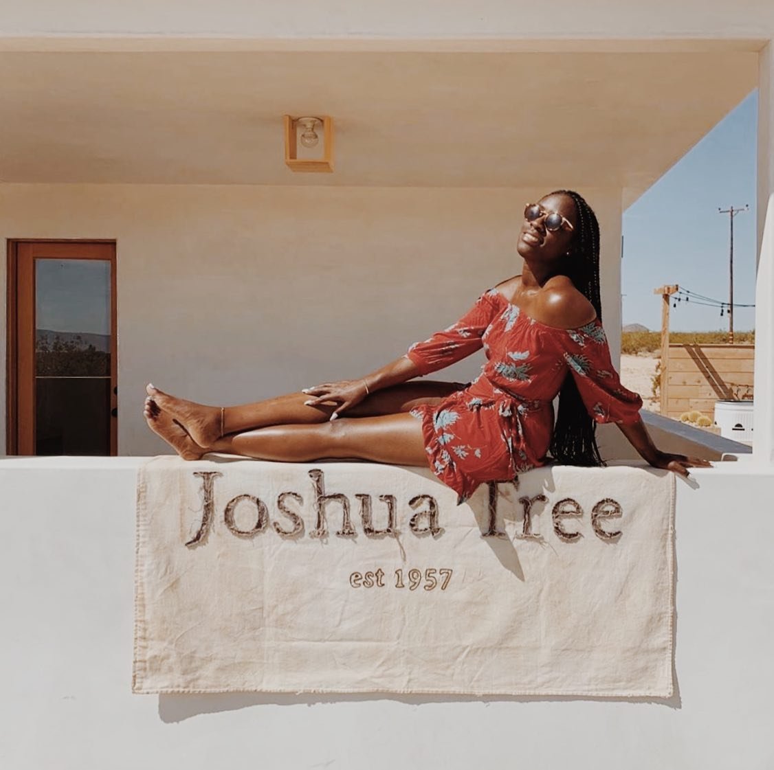 A woman in a red floral dress and sunglasses relaxing on a white flag with "Joshua Tree est 1957" embroidered on it, sitting outdoors on a ledge with a desert background.