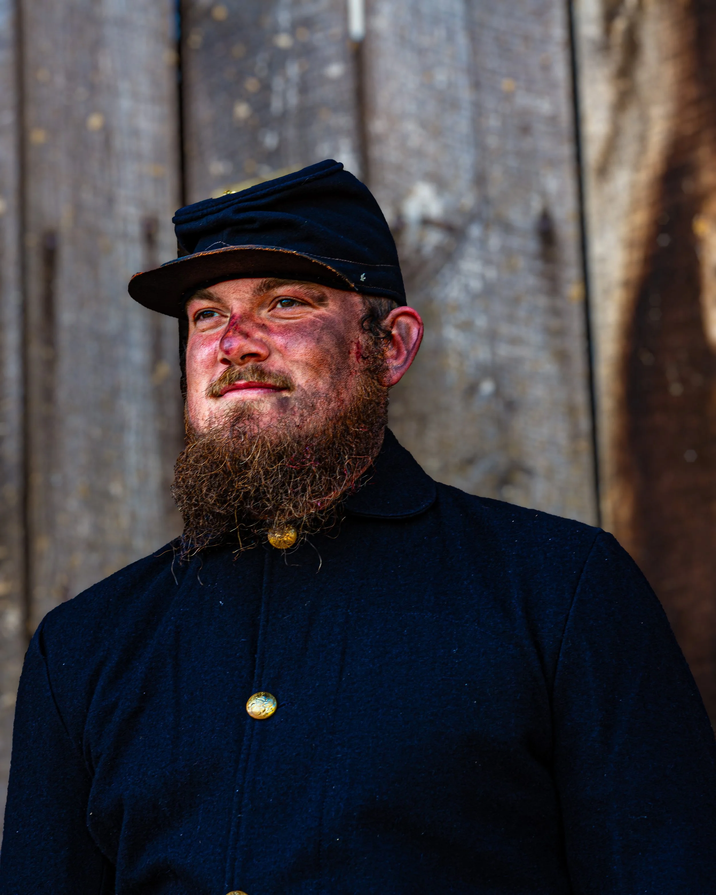 Close-up of a man with a beard and mustache, wearing a black cap and navy blue coat with gold buttons, standing in front of a weathered wooden fence.