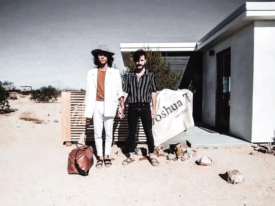 Two people standing in front of a modern house in a desert area, holding hands. The woman on the left is wearing a white blazer, tan top, white pants, and a wide-brimmed hat, with a brown bag on the ground beside her. The man on the right is dressed 