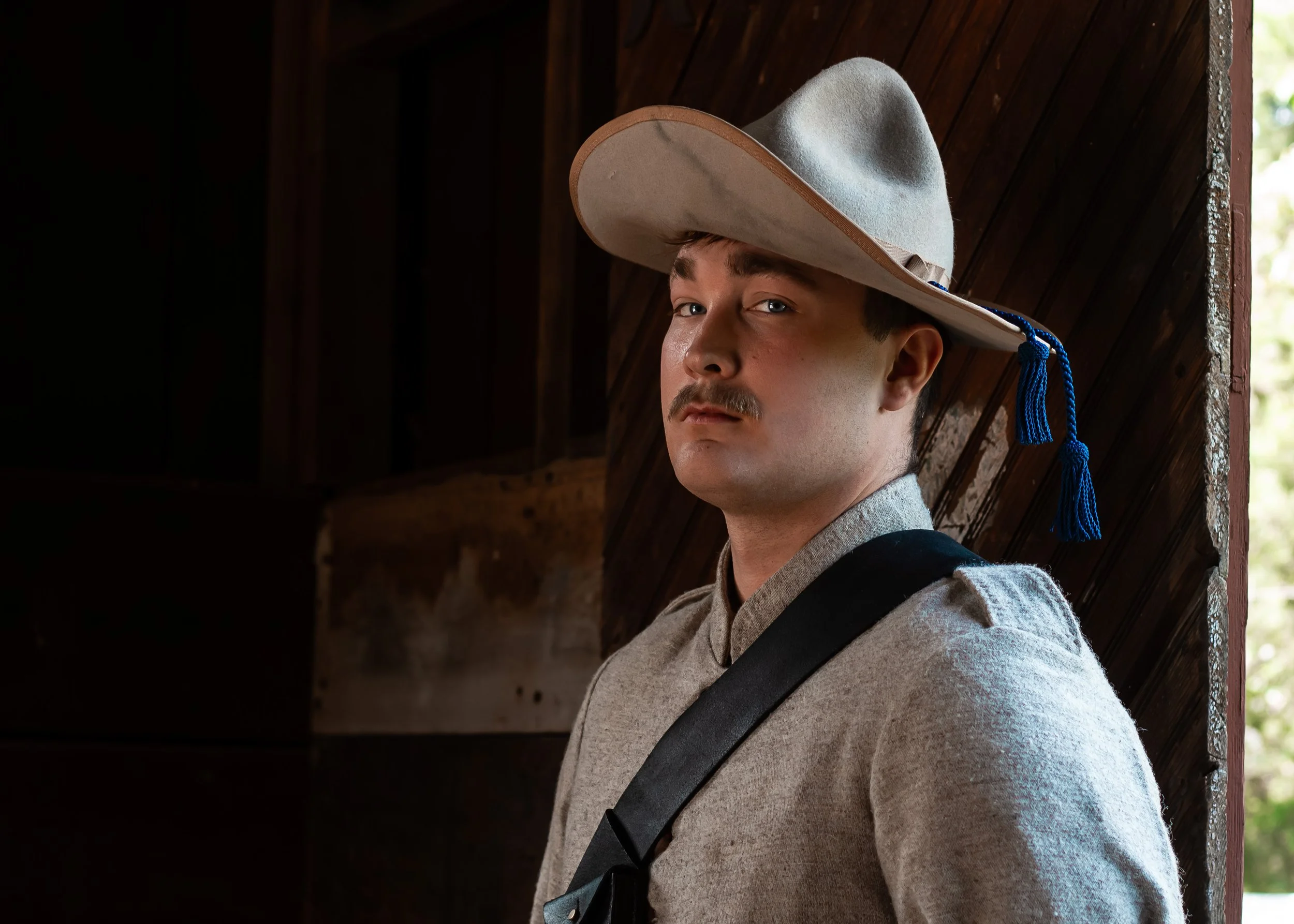 A young man with a cowboy hat, gray shirt, and black strap, standing indoors against a wooden wall. Light coming from the window on the right side.