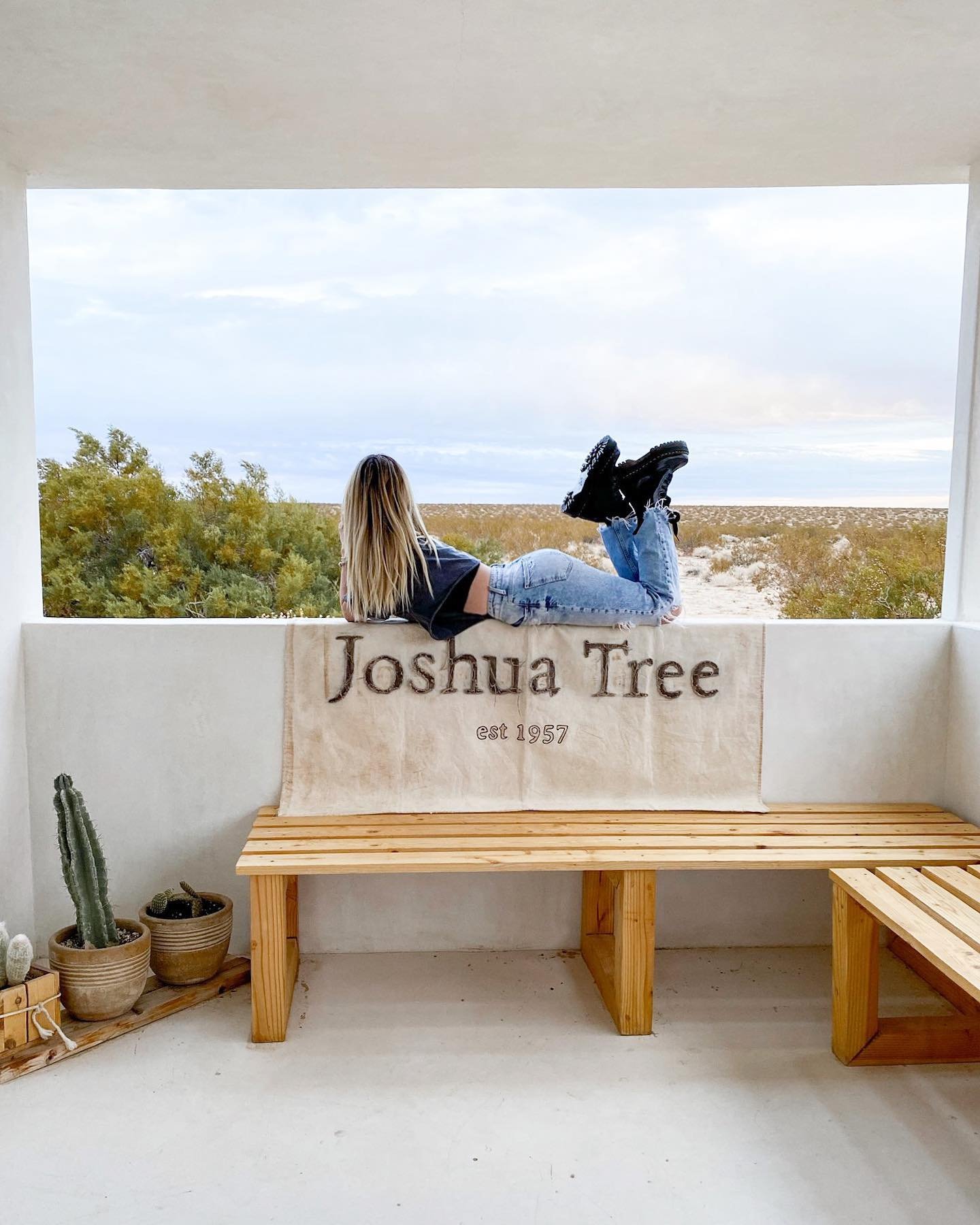 A woman with long blonde hair lying on a balcony ledge overlooking a desert landscape, wearing a black top, ripped jeans, and black shoes, with a flag that reads 'Joshua Tree est 1957'.