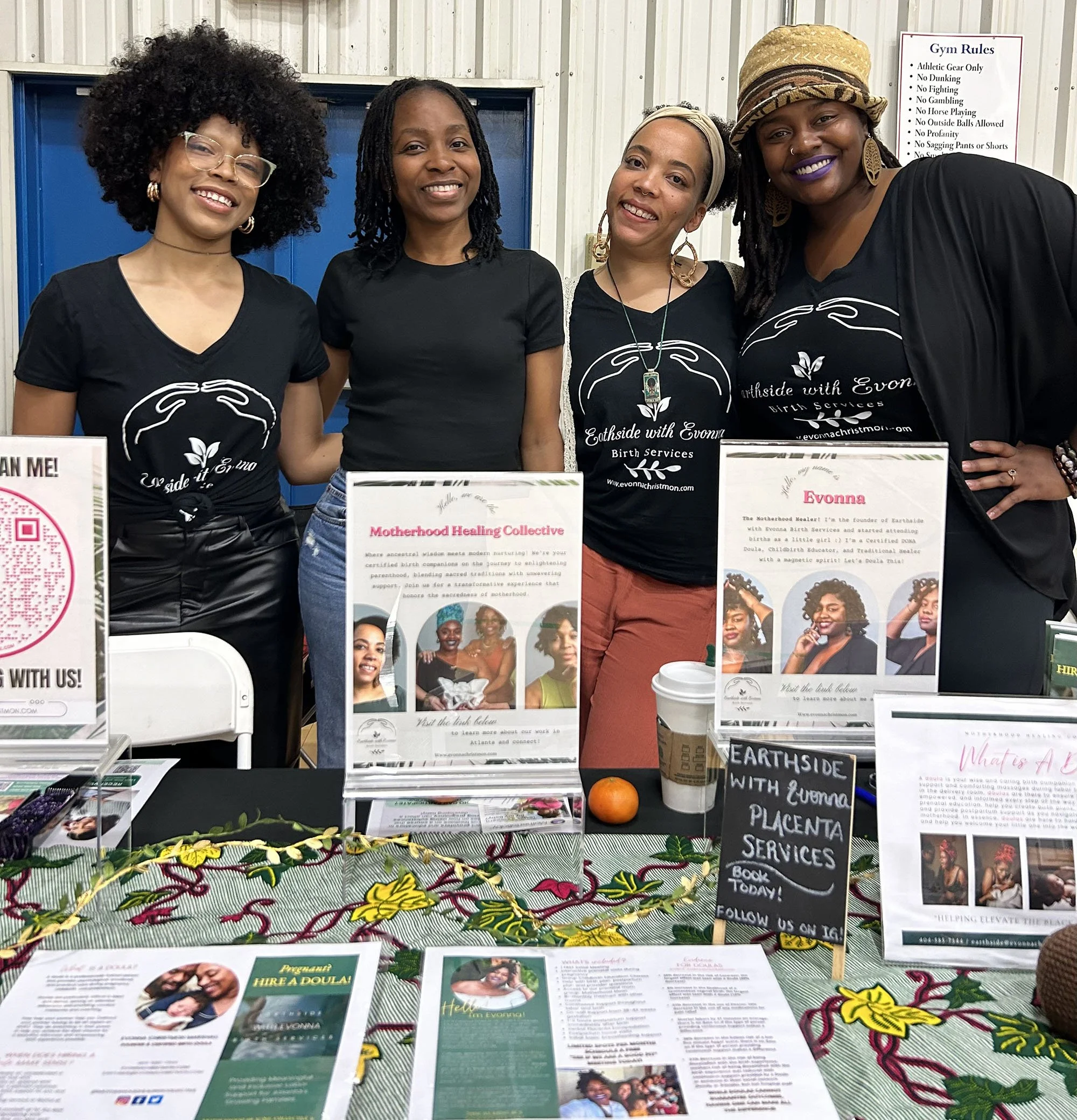 A group of four women standing behind a table at an event. The table displays informational materials and brochures related to birth services and wellness. The setup includes signs for "Motherhood Healing Collective" and "Earthside with Evonna Placenta Services." The women are smiling, and two of them are wearing matching t-shirts with the words "Earthside with Evonna Birth Services." A colorful cloth covers the table.