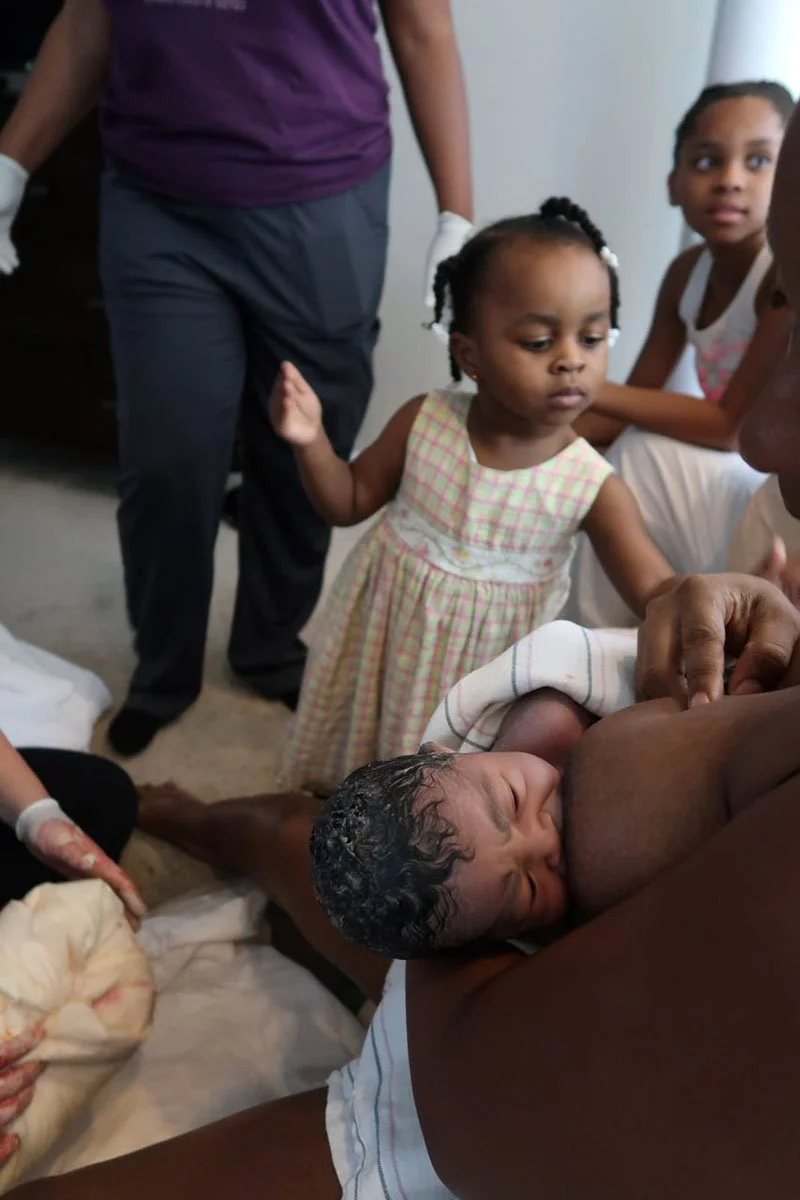 A mother breastfeeding a newborn baby while a young girl in a plaid dress observes, with other people nearby in a domestic setting.