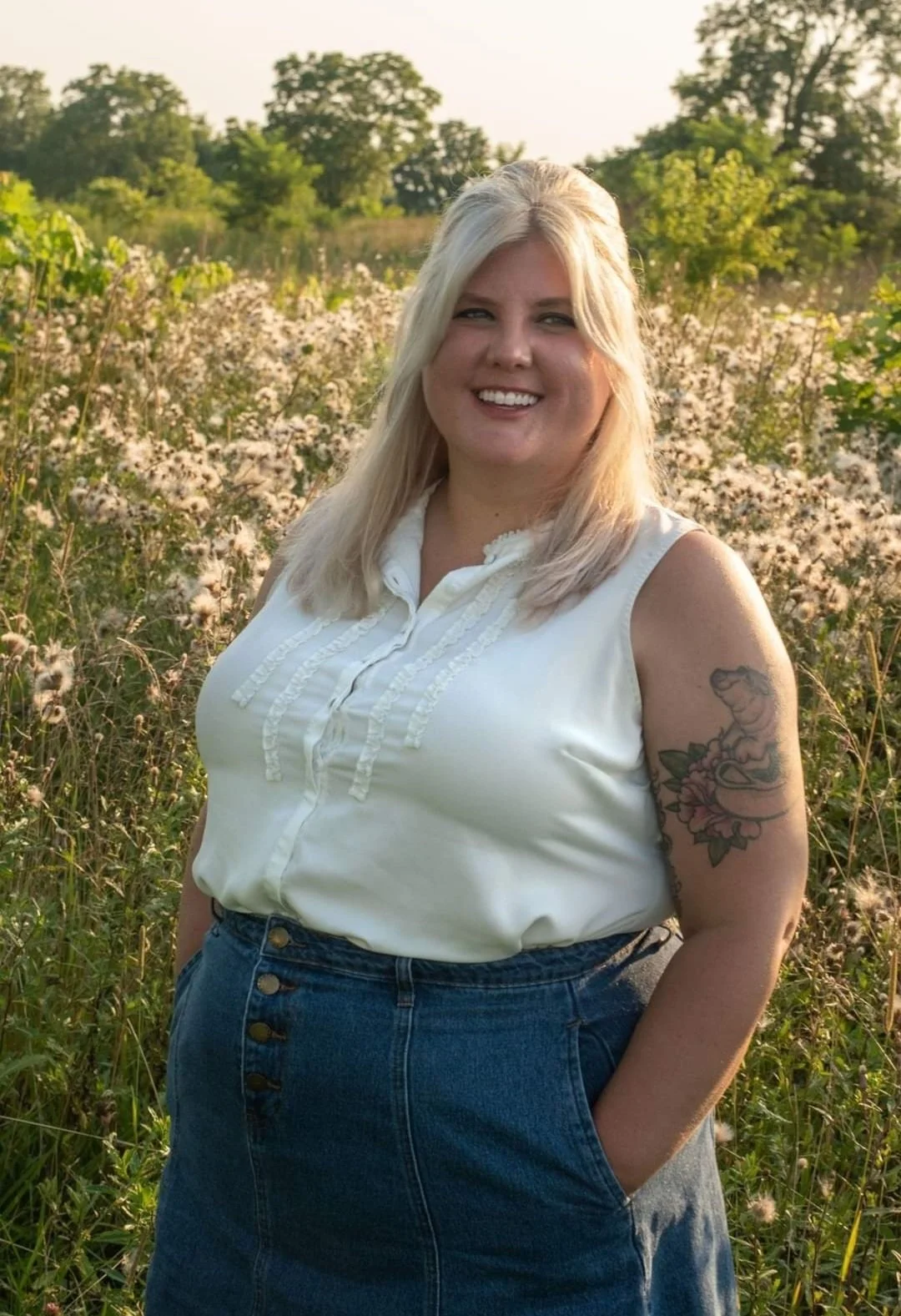 Person with long blonde hair wearing a white sleeveless blouse and a denim skirt stands in a field with wildflowers.