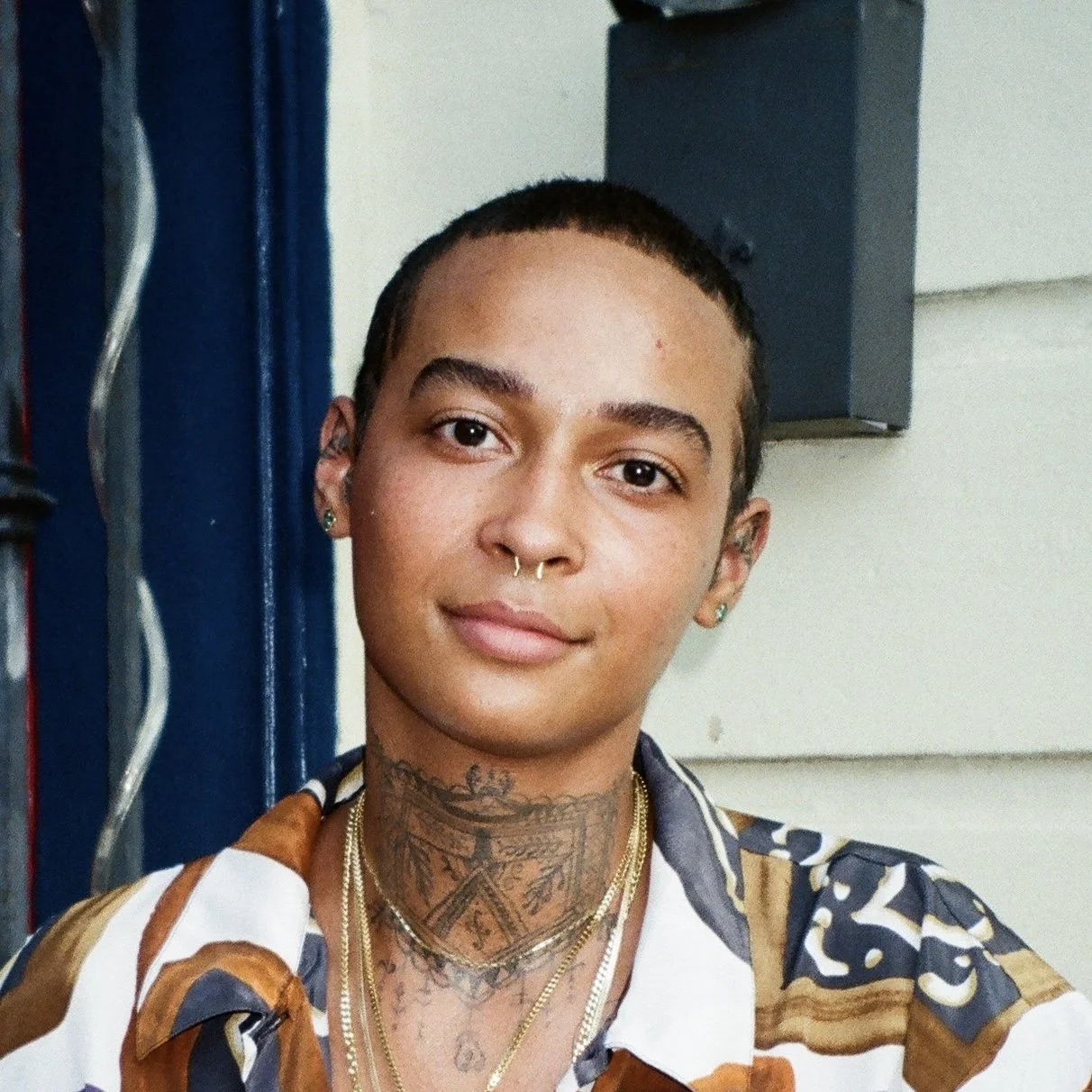 Person with short hair and tattoos, wearing a patterned shirt, posing for a portrait.