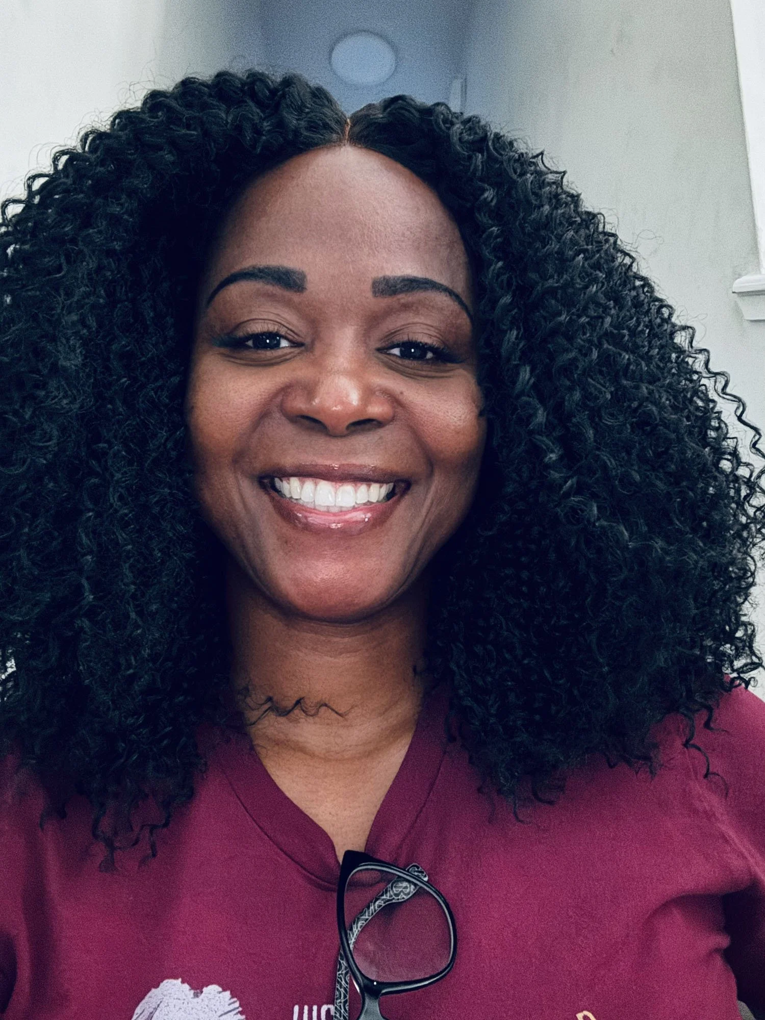 Woman smiling with curly black hair wearing a maroon shirt and glasses resting on the collar.