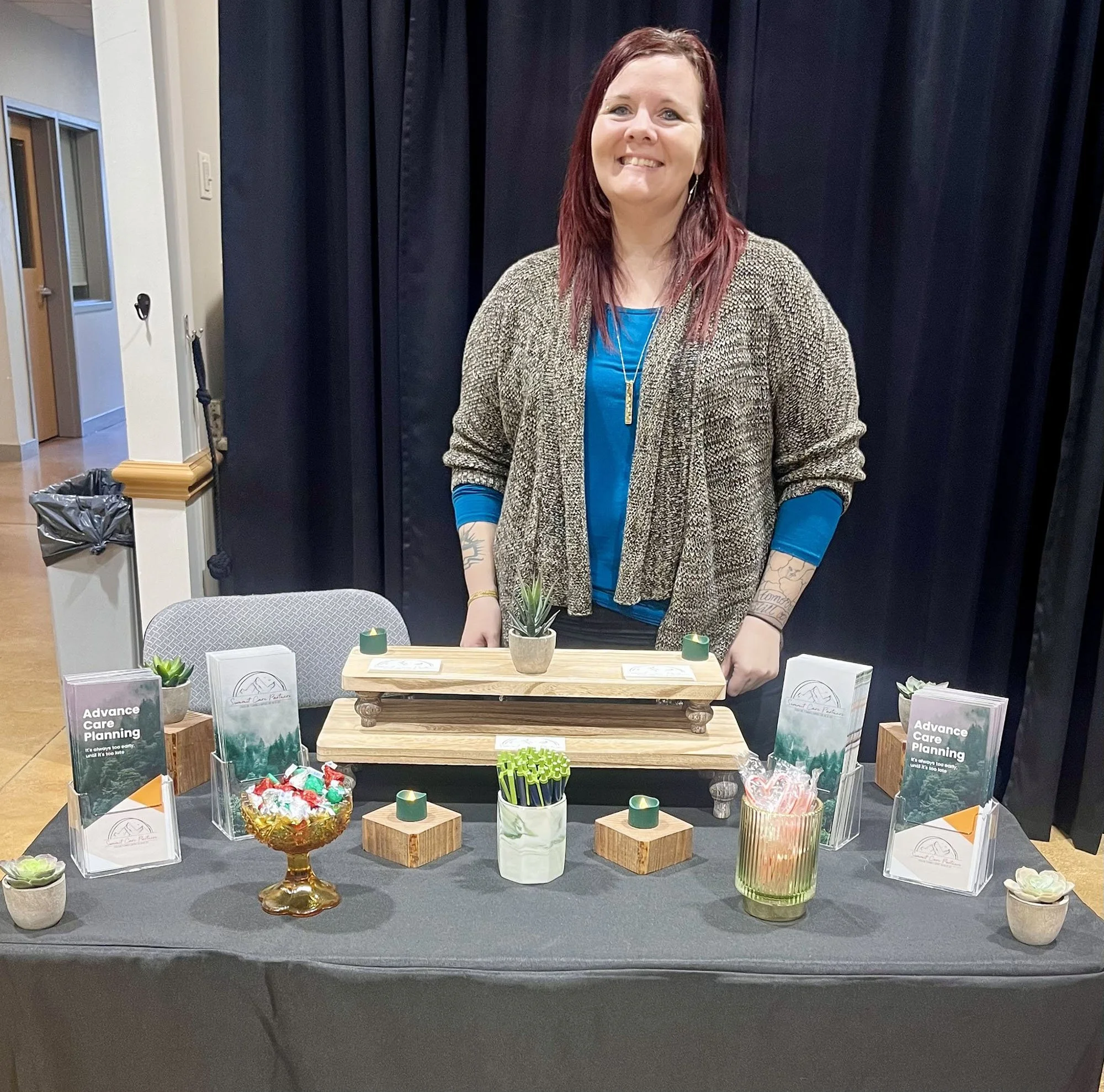 Woman at a display table with brochures titled 'Advance Care Planning', small potted plants, candy, and pens in a decorated setup.