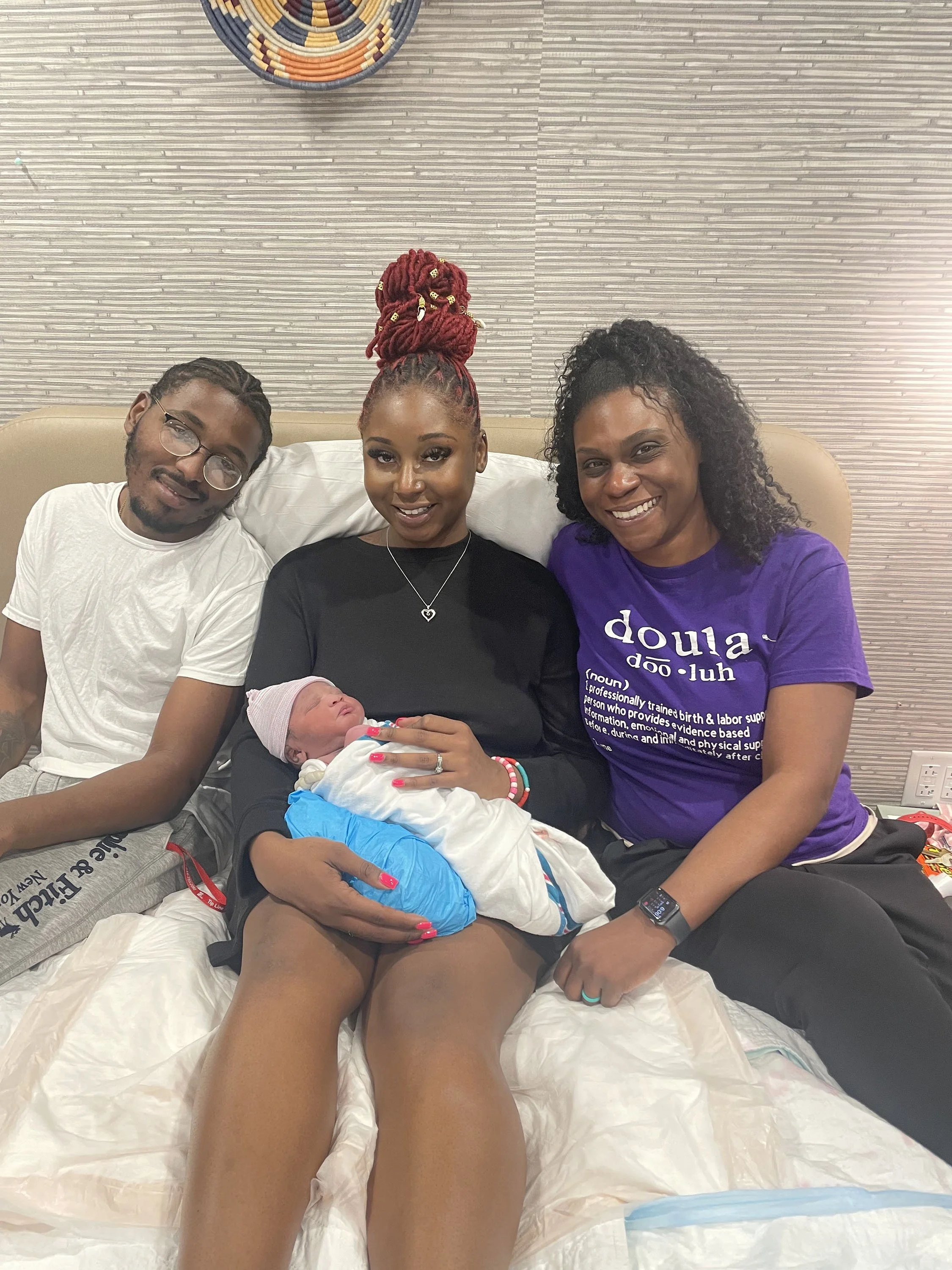 Three adults smiling with a newborn baby, seated indoors. One of the adults is wearing a purple "doula" t-shirt, and the baby is wrapped in white and blue blankets.