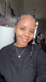 A person smiling indoors, wearing a black sweater and a necklace, with braided hair.