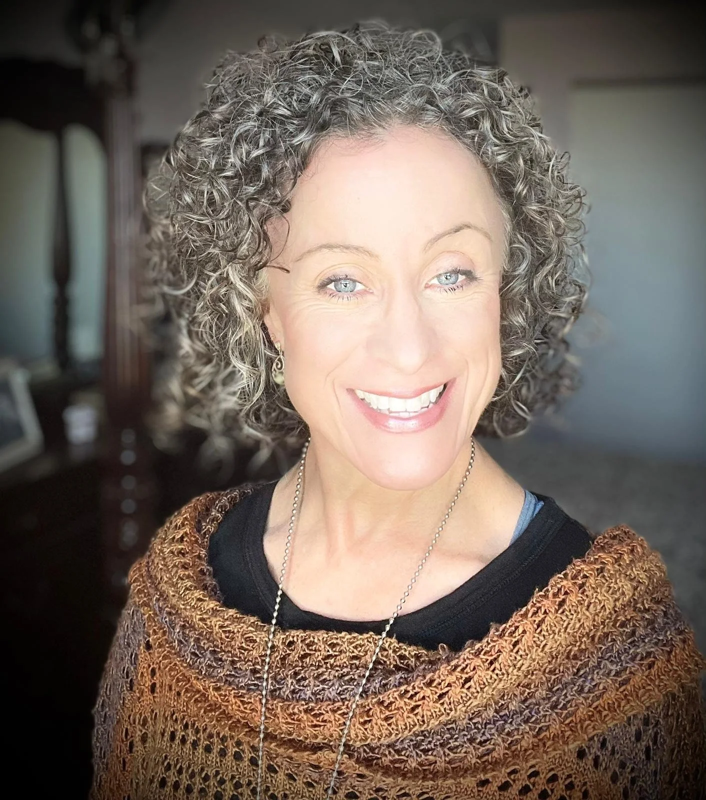 Smiling woman with curly gray hair and a crocheted top in a softly lit room.