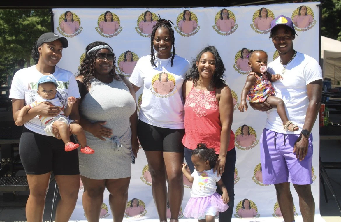 Group of six people, including two children, posing in front of a backdrop with a repeated design. Some adults are holding the children, and one adult is wearing a graphic shirt. The backdrop features a logo depicting a person and text. Everyone is smiling.