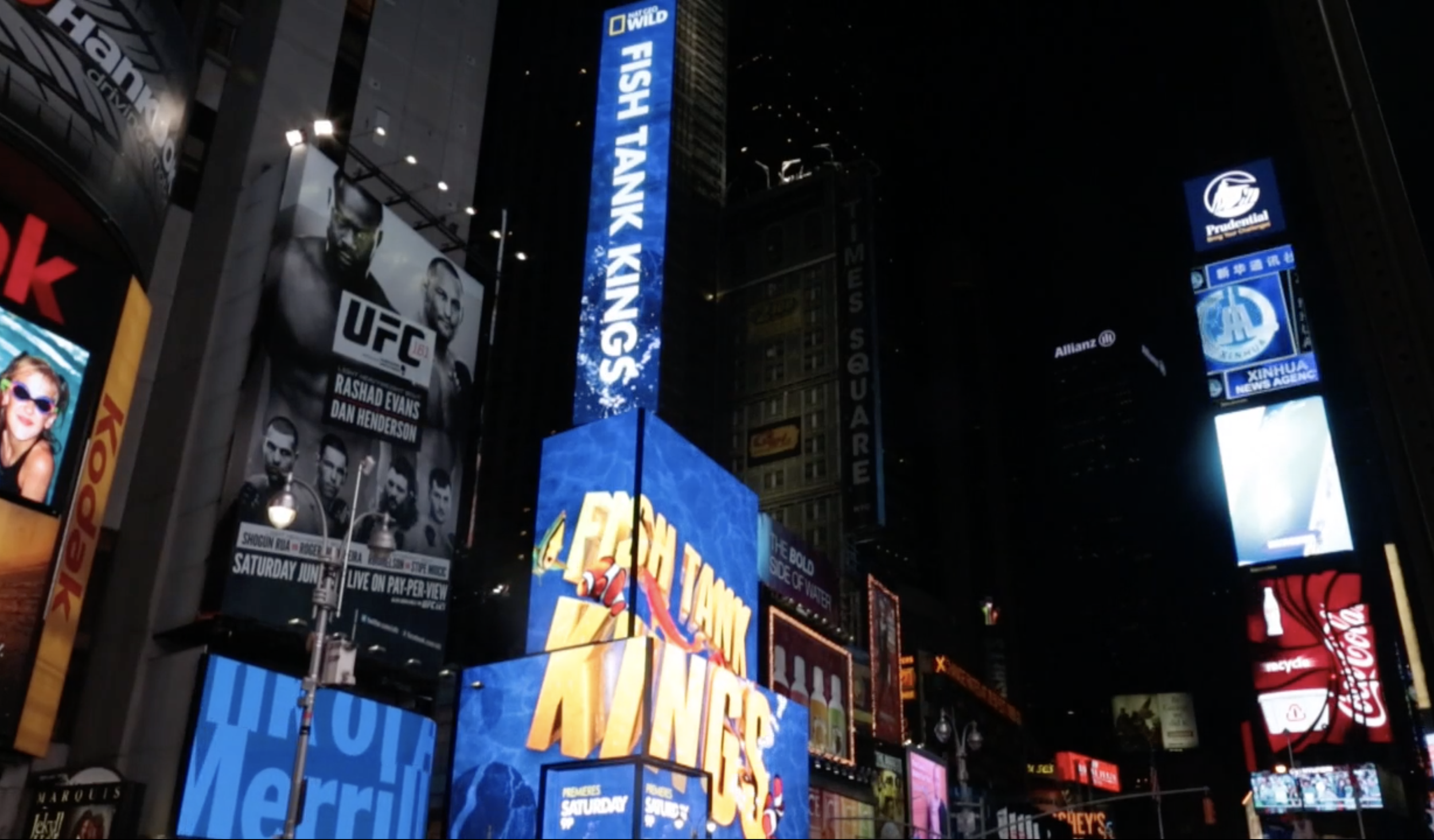 Nighttime scene in Times Square, New York City, with bright digital billboards displaying advertisements, including one for UFC and another for Yas Queen, and various neon signs and logos.