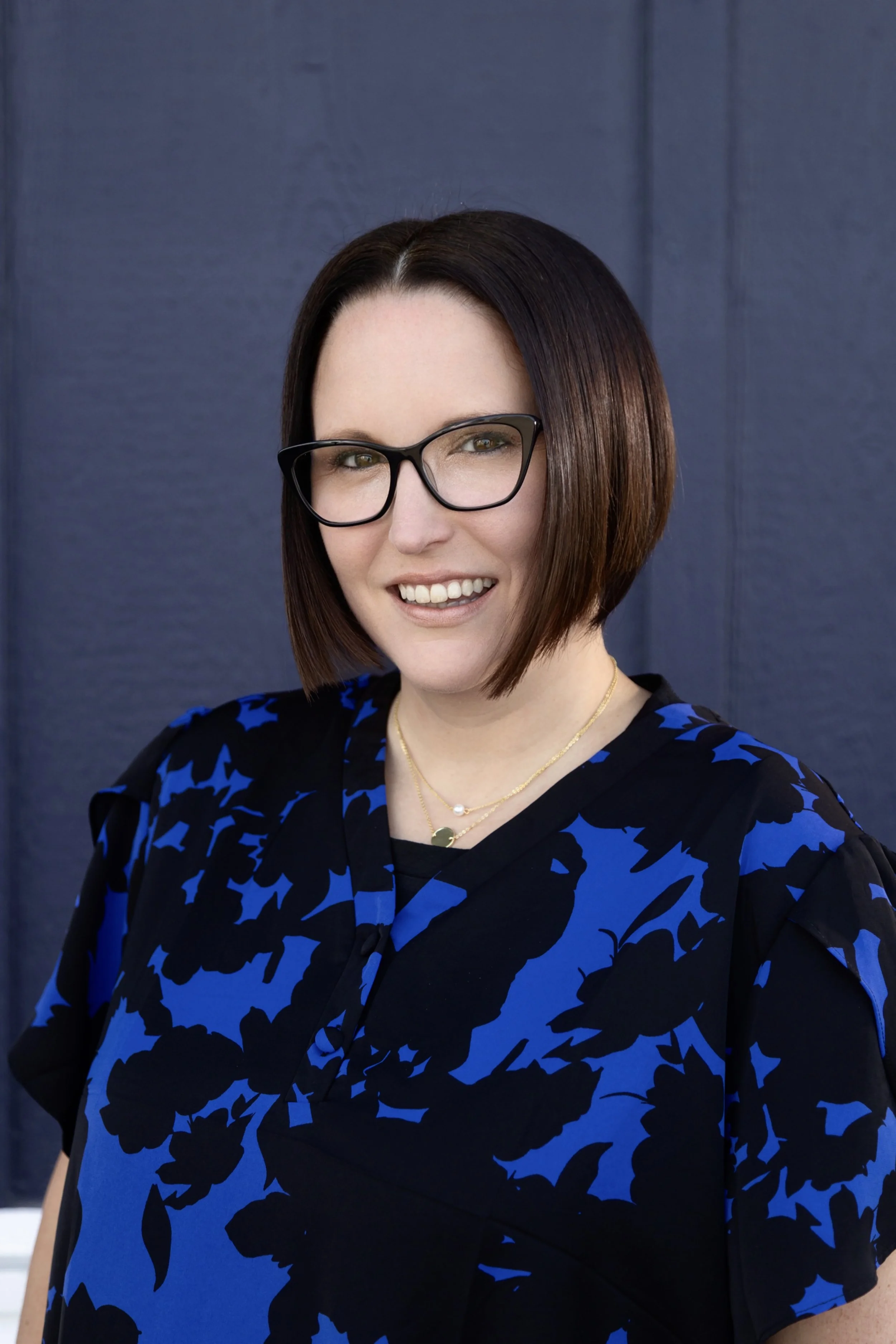 A young woman with shoulder-length dark brown hair, glasses, and a floral-patterned gray jacket, smiling outdoors with trees and greenery in the background.