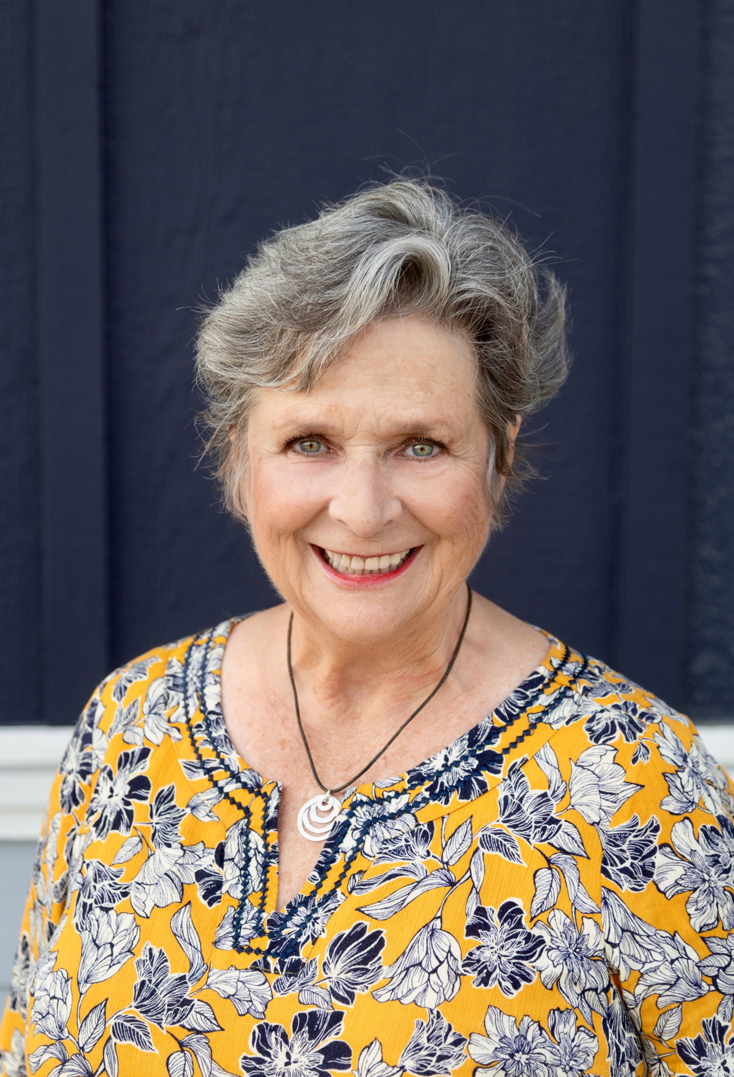 A woman with glasses and curly hair wearing a yellow floral top and a black necklace with a pendant, smiling, in front of a white horizontal panel background.