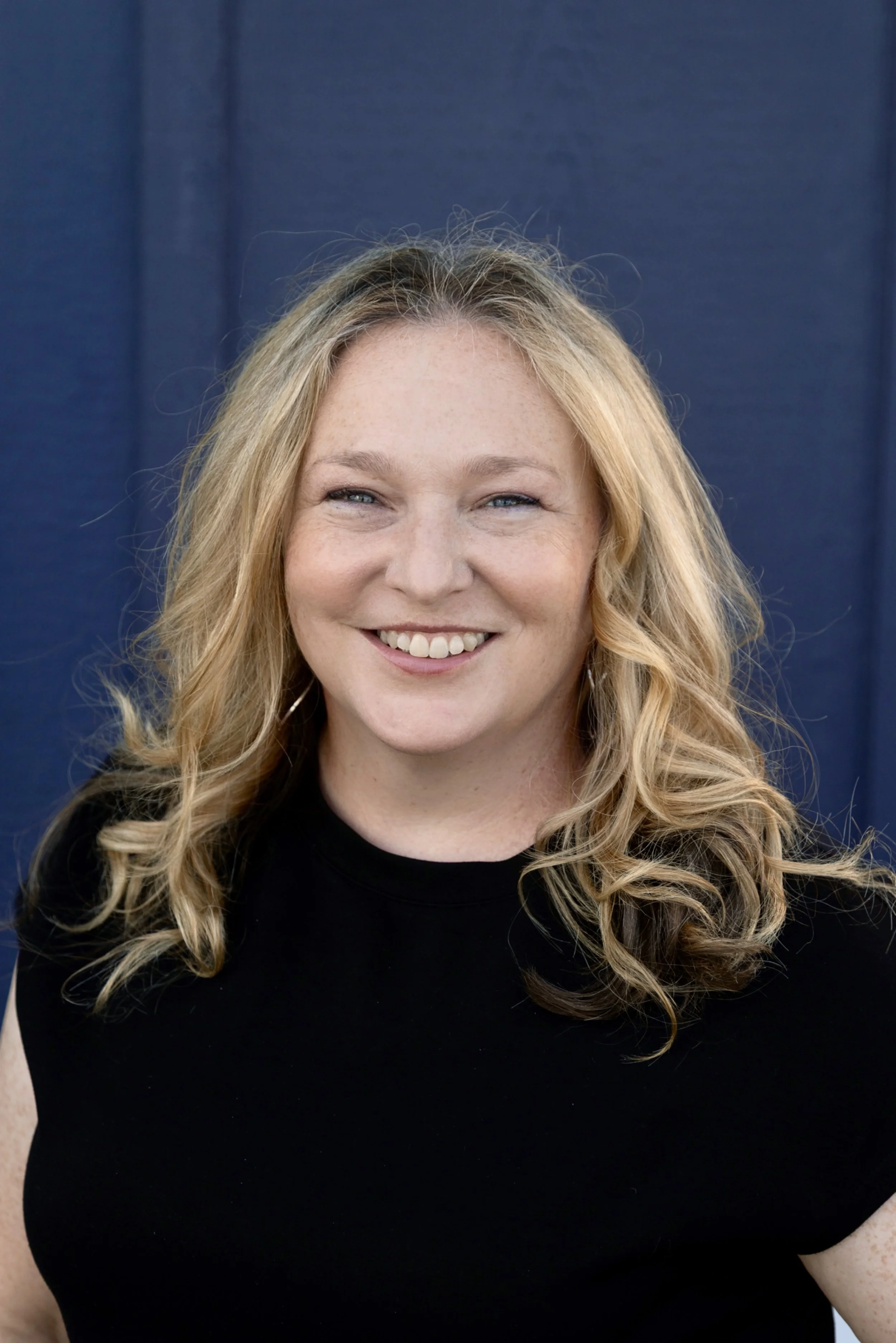 Portrait of a woman with long blonde hair and a black top smiling against a white paneled background.