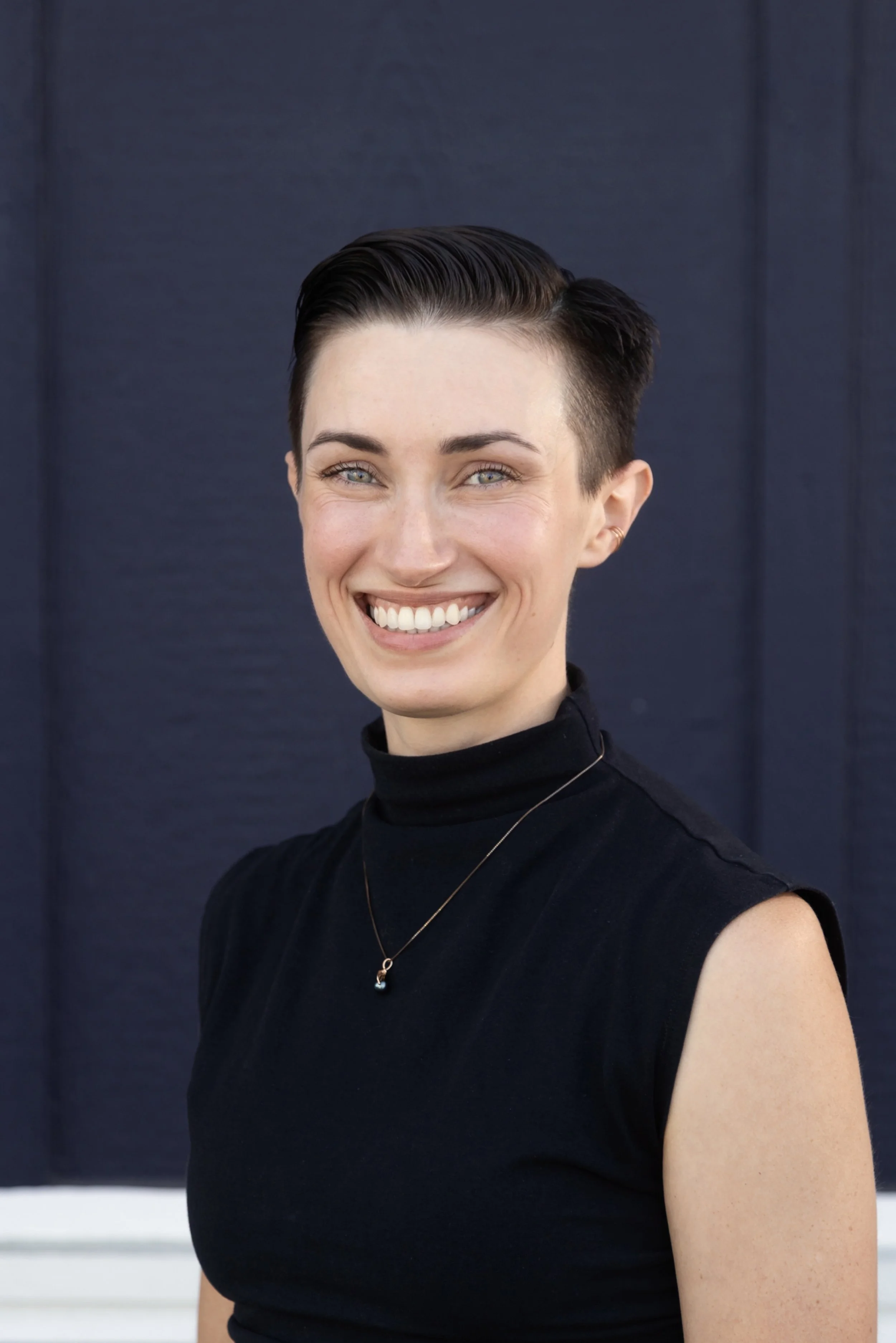 A smiling woman with short brown hair wearing a green scarf and a light gray speckled shirt standing against a white horizontal panel wall.