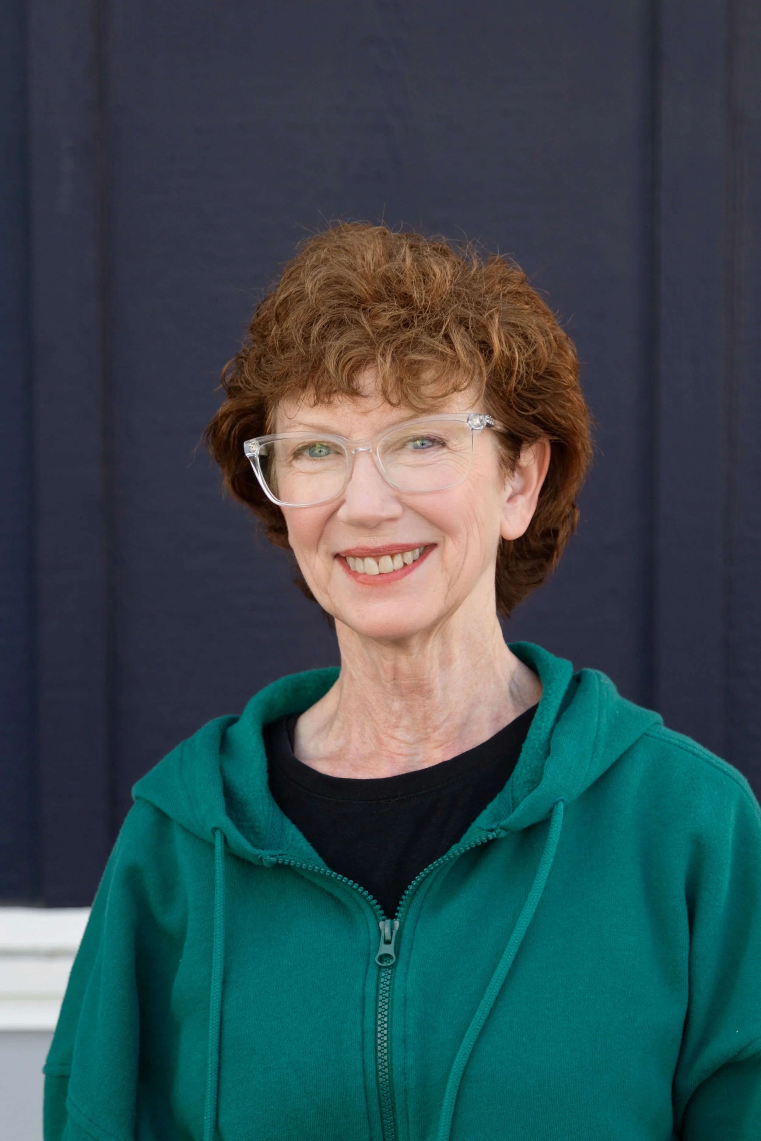 A woman with short, curly red hair smiling and wearing a light blue sweater standing against a white horizontal siding background.