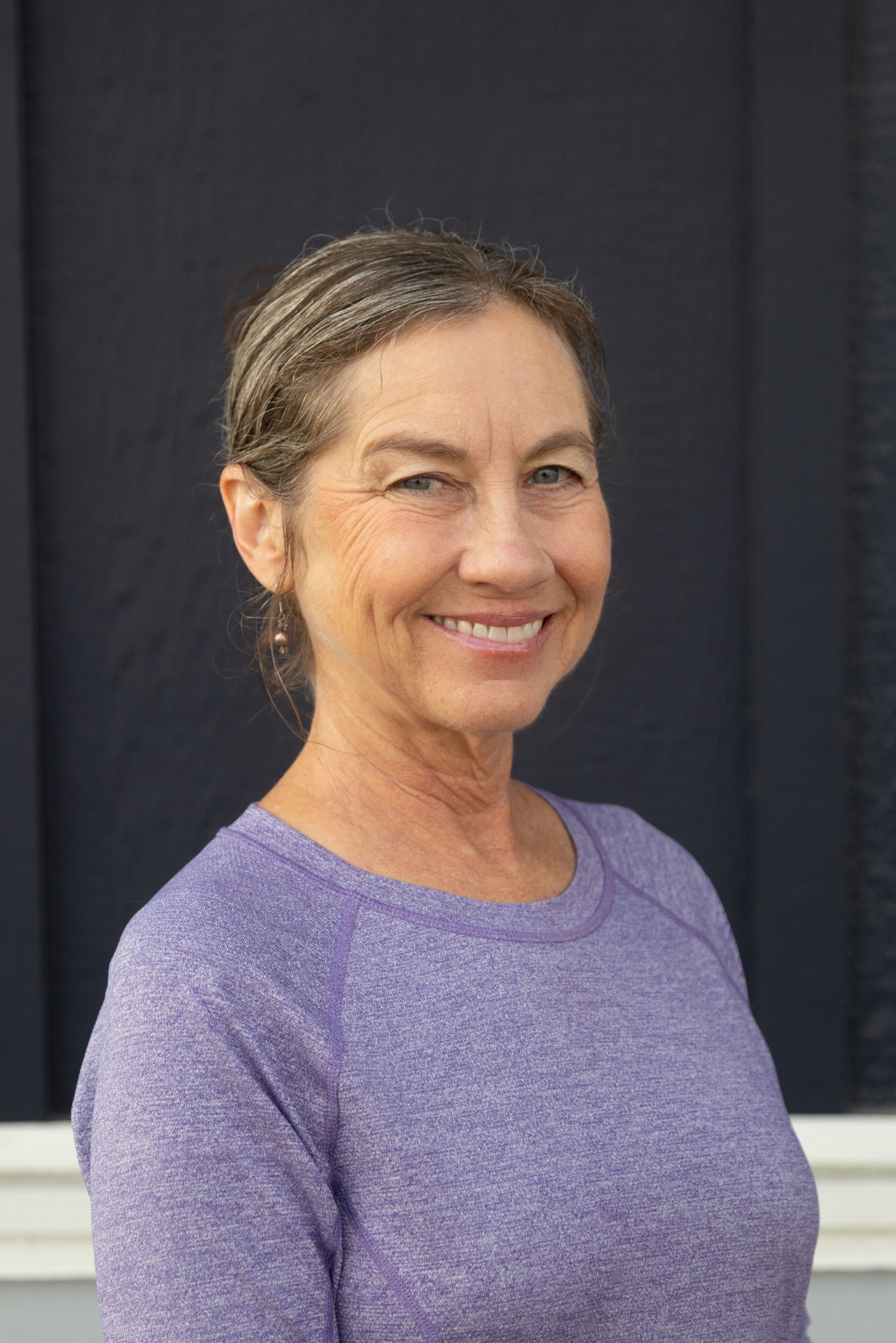 A smiling woman with short curly brown hair, wearing a black shirt, against a blurred neutral background.
