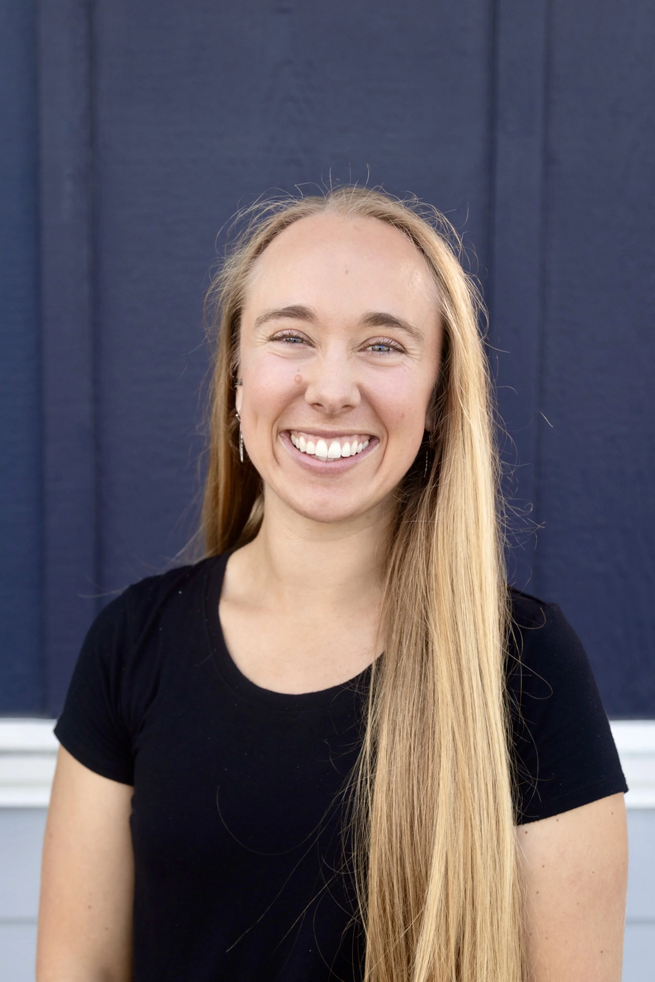 A smiling young woman with shoulder-length strawberry blonde hair, wearing a burgundy top, standing in front of a white horizontal-paneled wall.