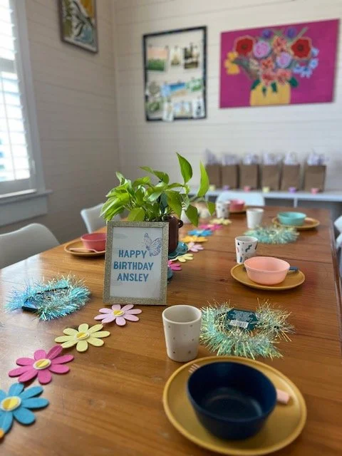 A table decorated with colorful paper flowers, shiny tinsel, and small cups and bowls, with a birthday sign reading 'Happy Birthday Ansley' in front of a potted plant. The room has wall art and bookshelves in the background.