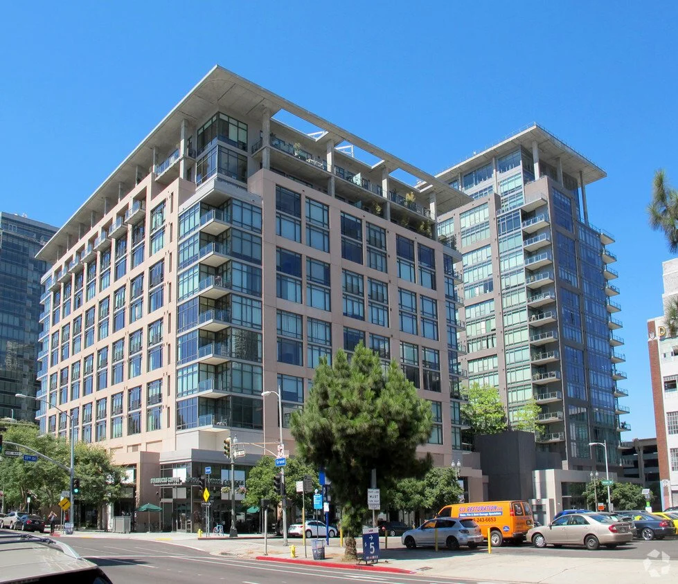 Modern multi-story glass and concrete residential or office building on a city street with cars and pedestrians, under a clear blue sky.