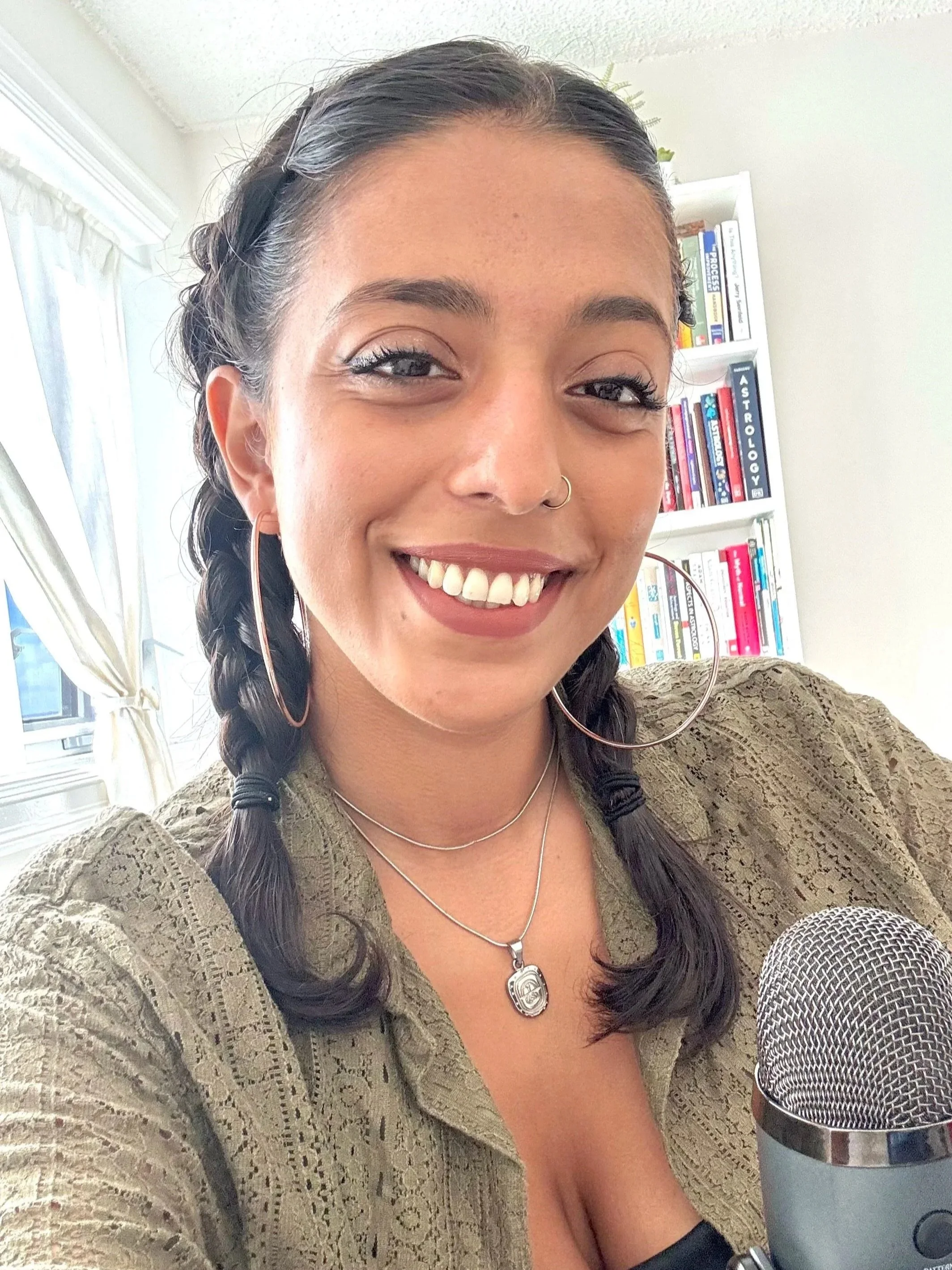 A woman with braided hair smiling at the camera, wearing hoop earrings, a necklace, and a green jacket, in front of a bookshelf filled with books and a window with curtains.