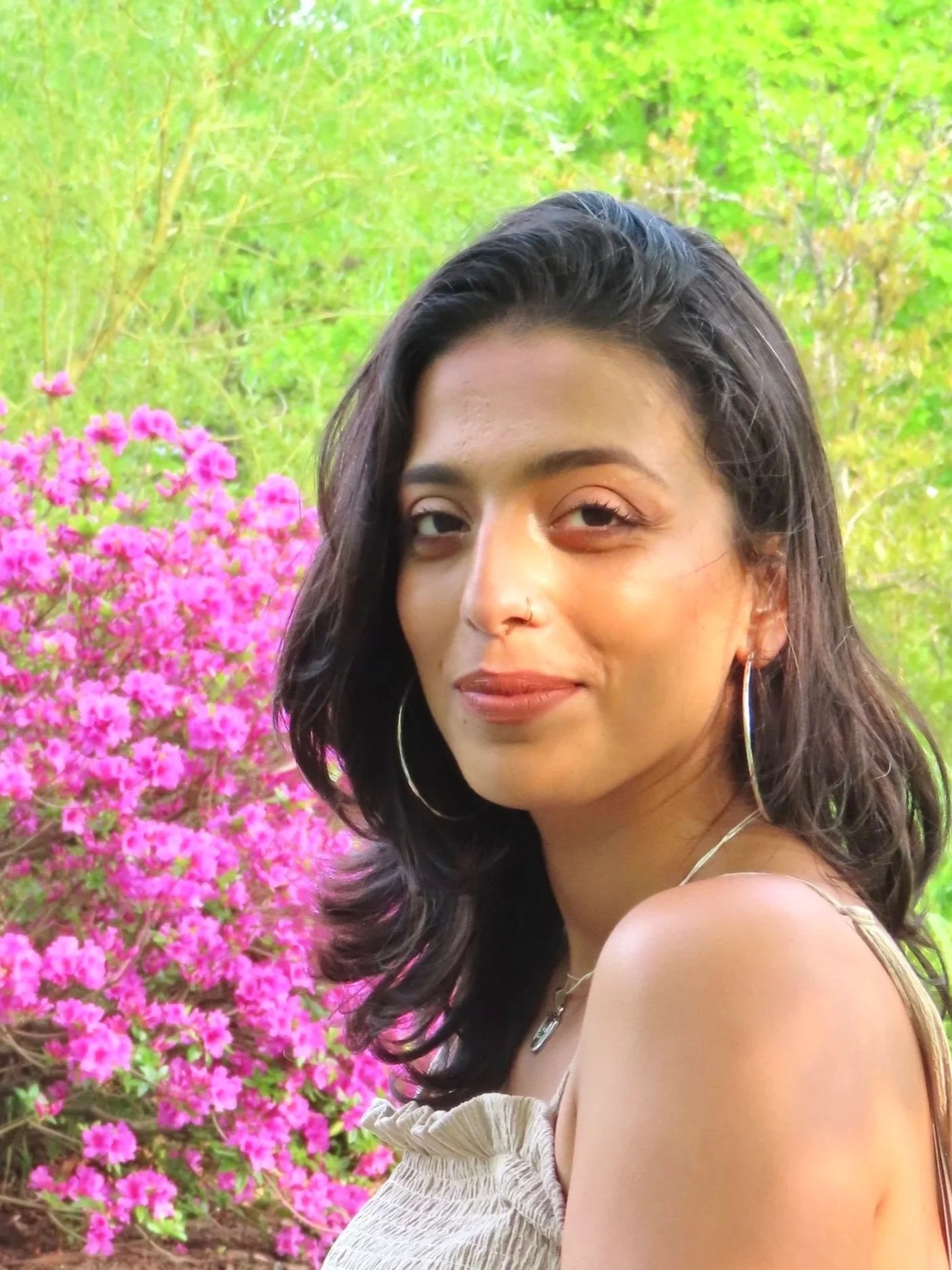 A woman with dark wavy hair and hoop earrings is smiling at the camera, standing outdoors with pink flowers and green foliage in the background.