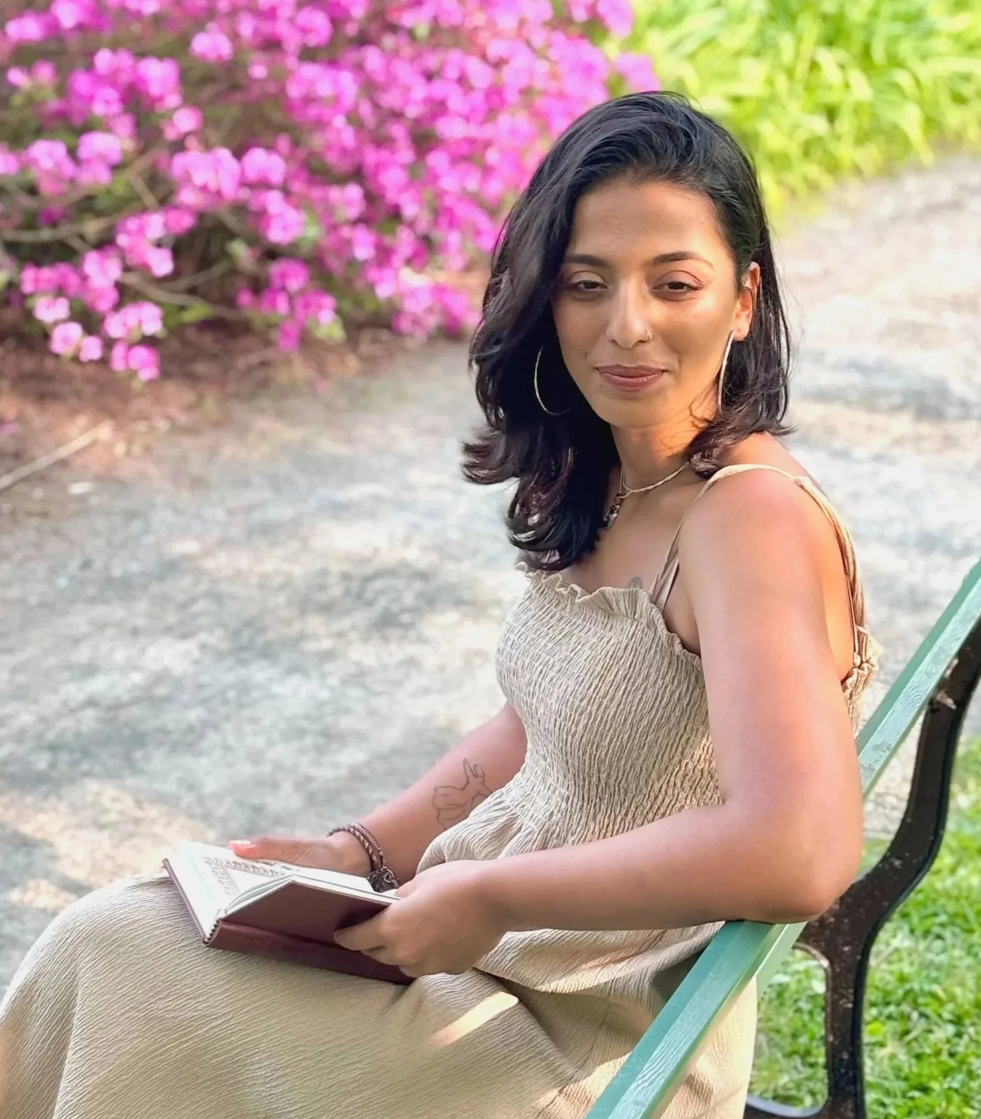 A woman sitting outdoors on a green park bench, holding an open book, with pink flowering bushes in the background.