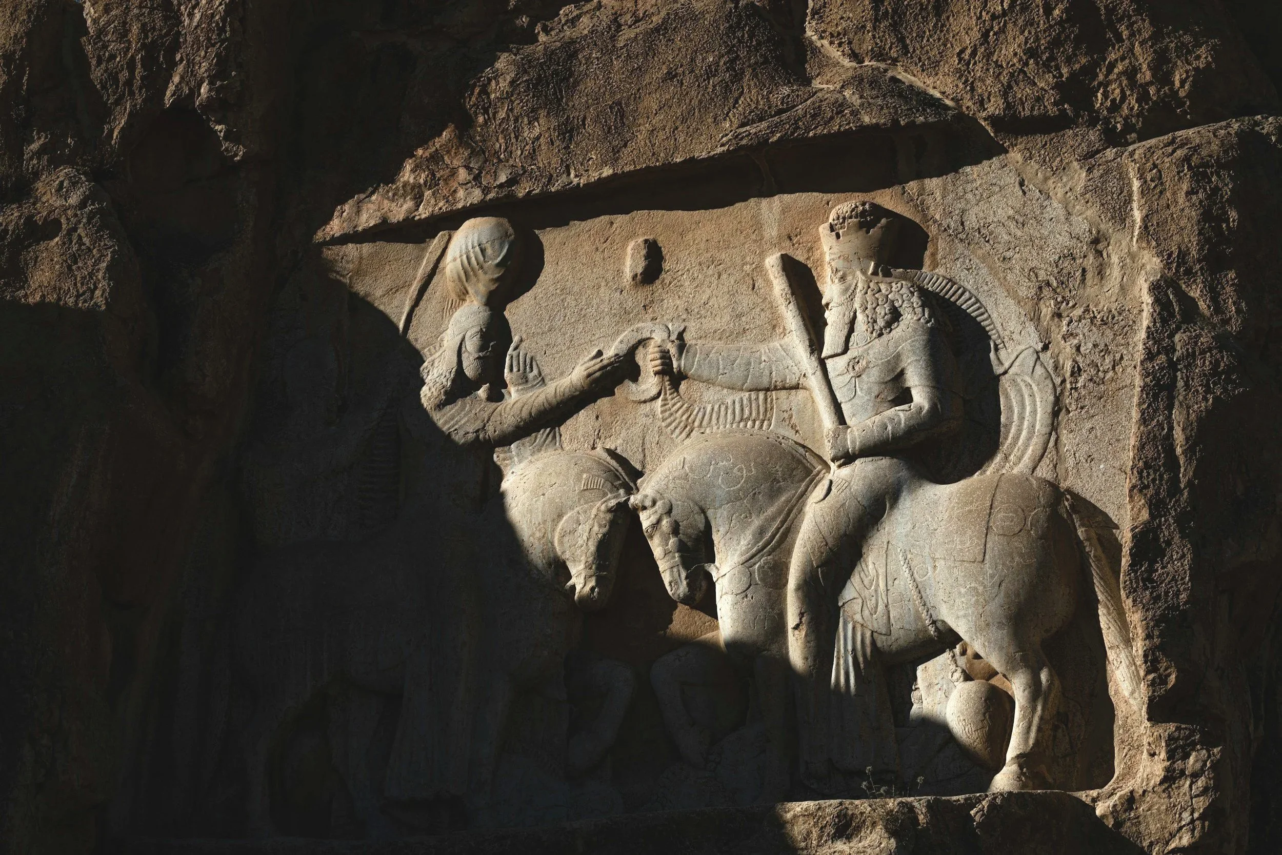 Ancient stone relief depicting mythological figures, including a winged creature and a regal figure wearing a crown, carved into a rock face.