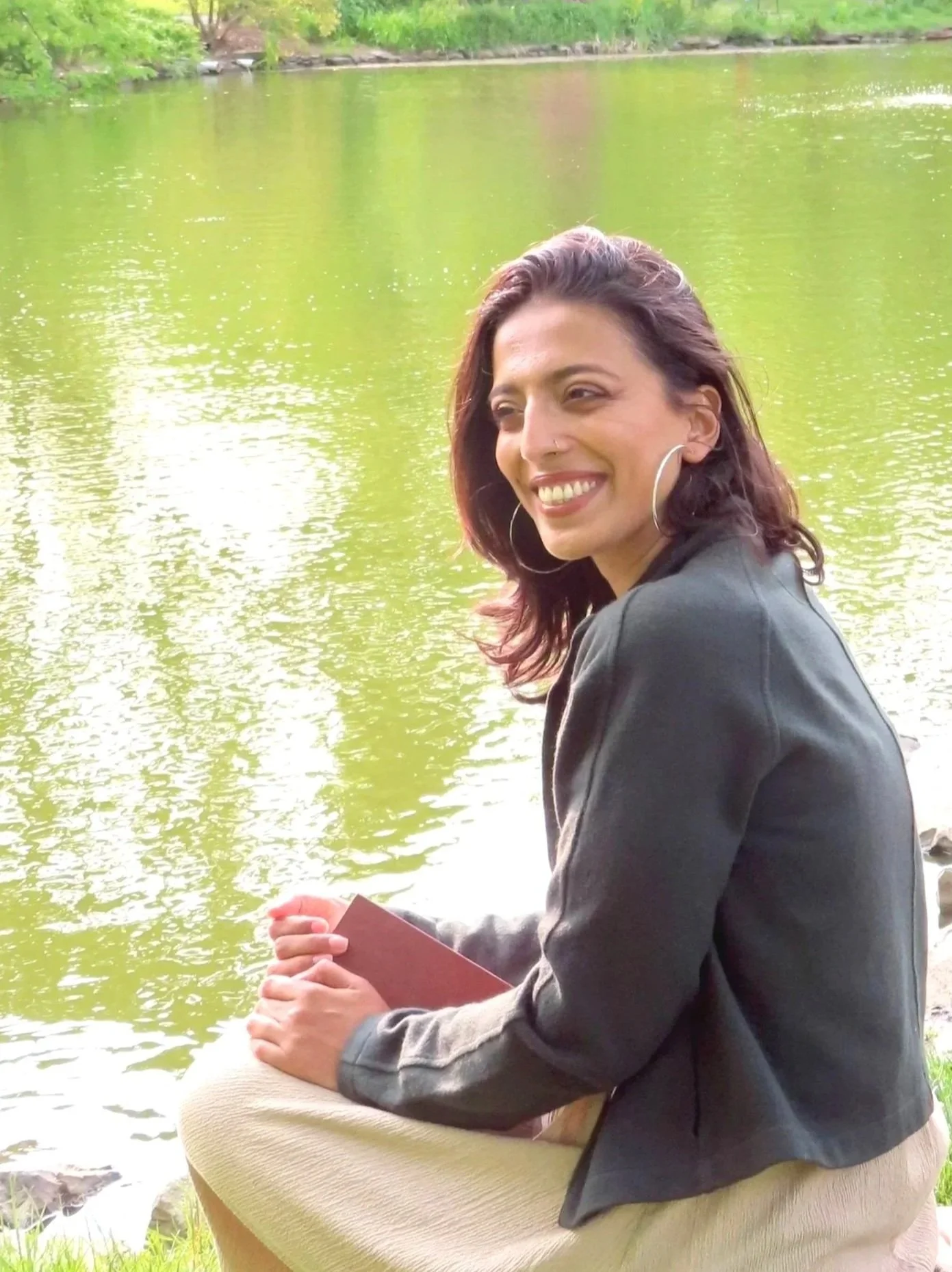 A woman sitting outdoors by a lake, smiling and holding a tablet, with lush greenery in the background.